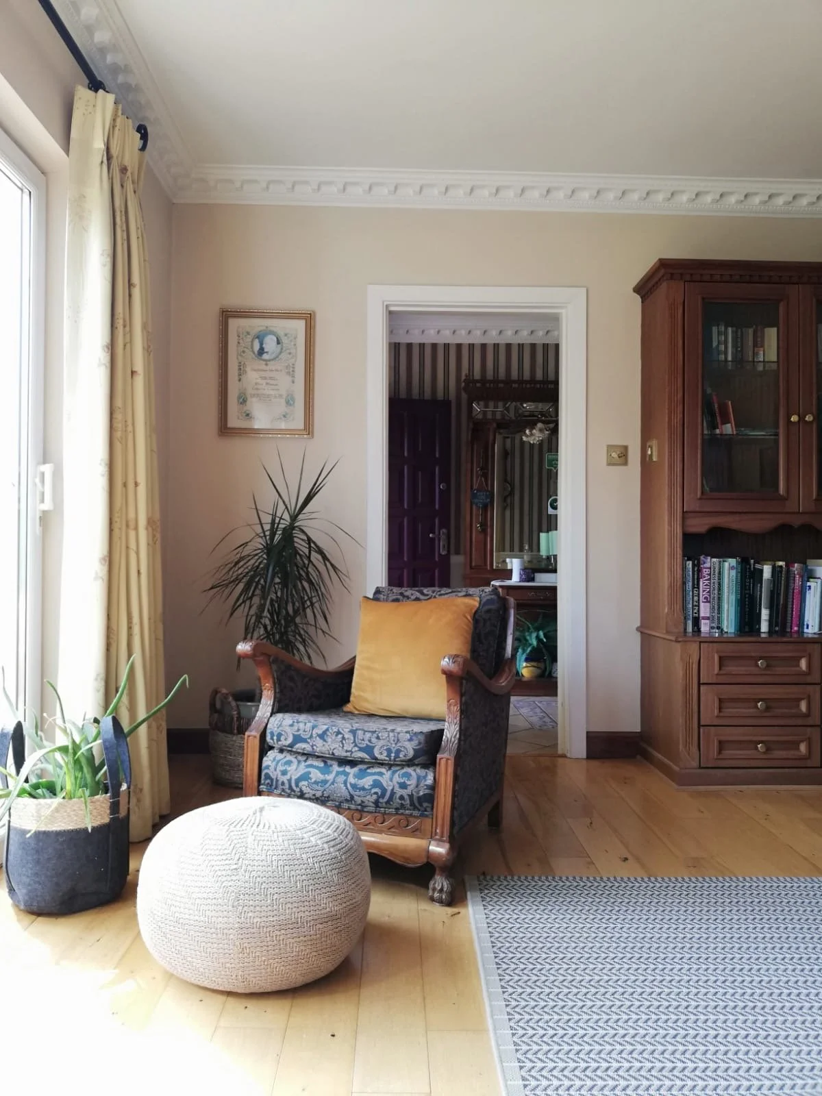 Living room with an armchair, a yellow pillow, a patterned pouf, tall plant, bookcase, doorway, and window with cream curtains.