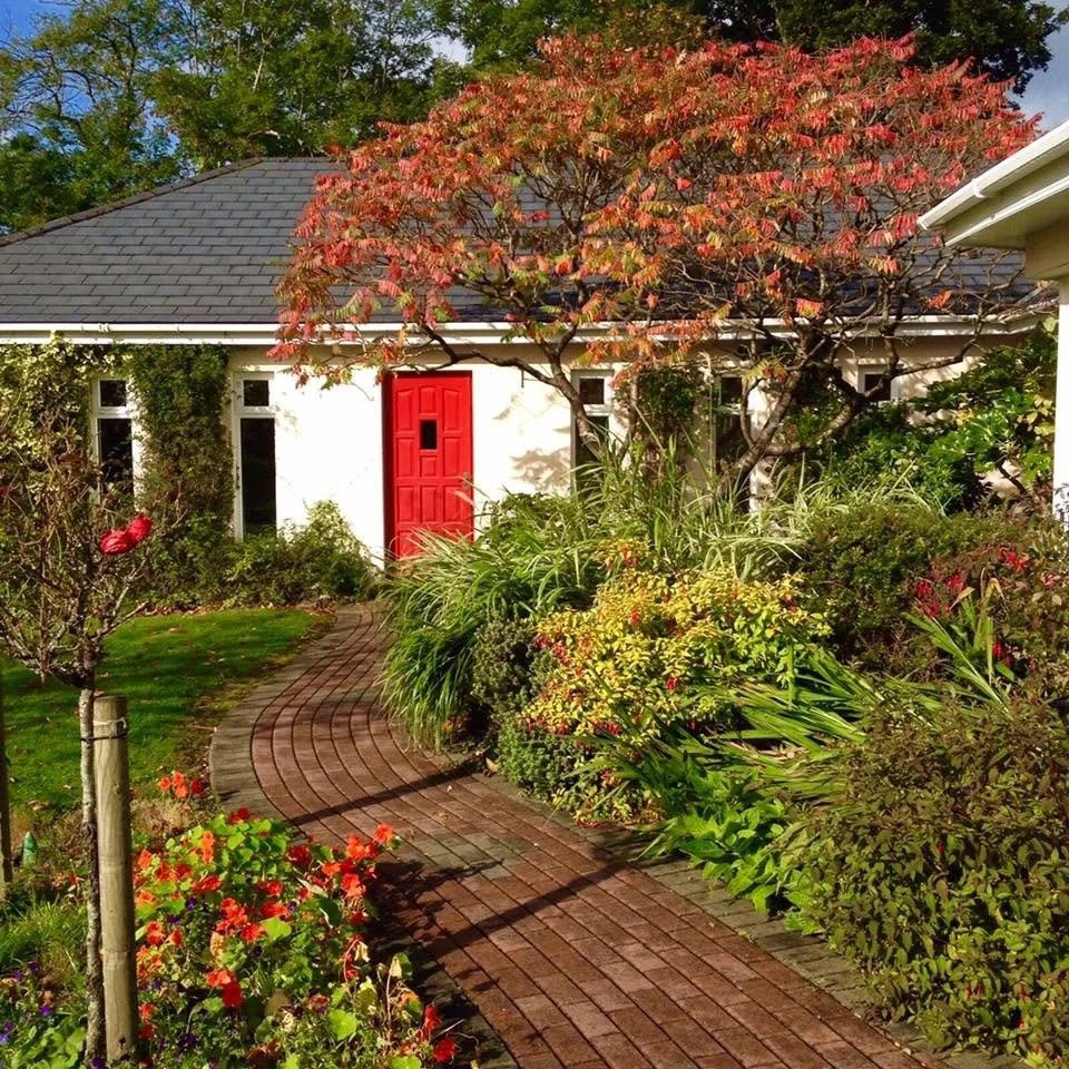 A garden path with red brick pavers winding through lush green plants and vibrant flowers leading to a white house with a red front door, a small tree with reddish-orange leaves, and surrounding trees and shrubs.