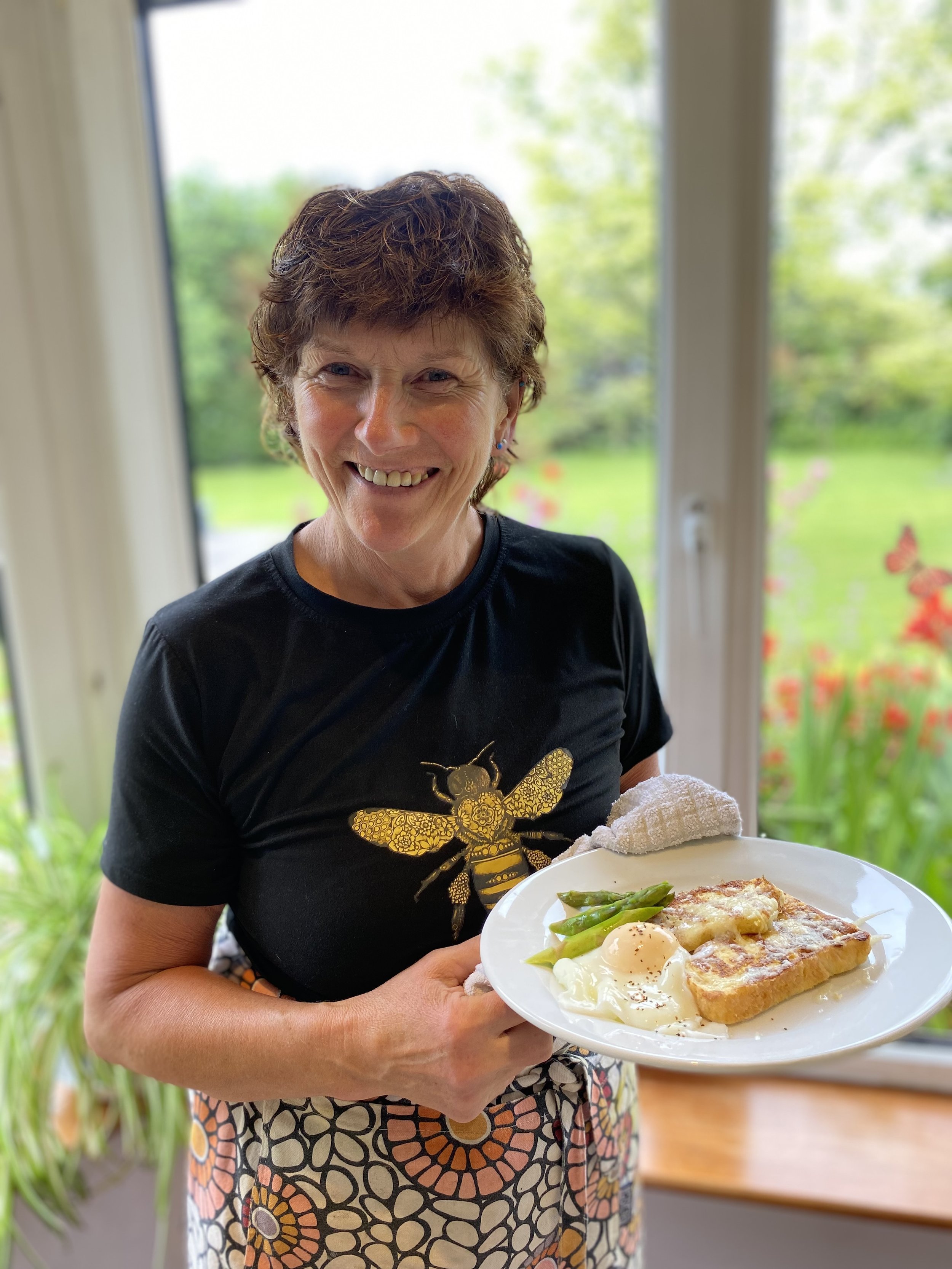 Smiling woman with short curly brown hair holding a plate of breakfast foods, including eggs, asparagus, and toast, standing near a large window with a garden view.