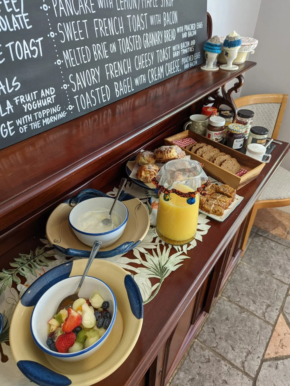 A breakfast spread on a wooden sideboard includes bowls of fruit, a pitcher of orange juice covered with a decorative cloth, slices of bread, muffins, yogurt, and various jars of preserves. There is also a chalkboard menu in the background listing br