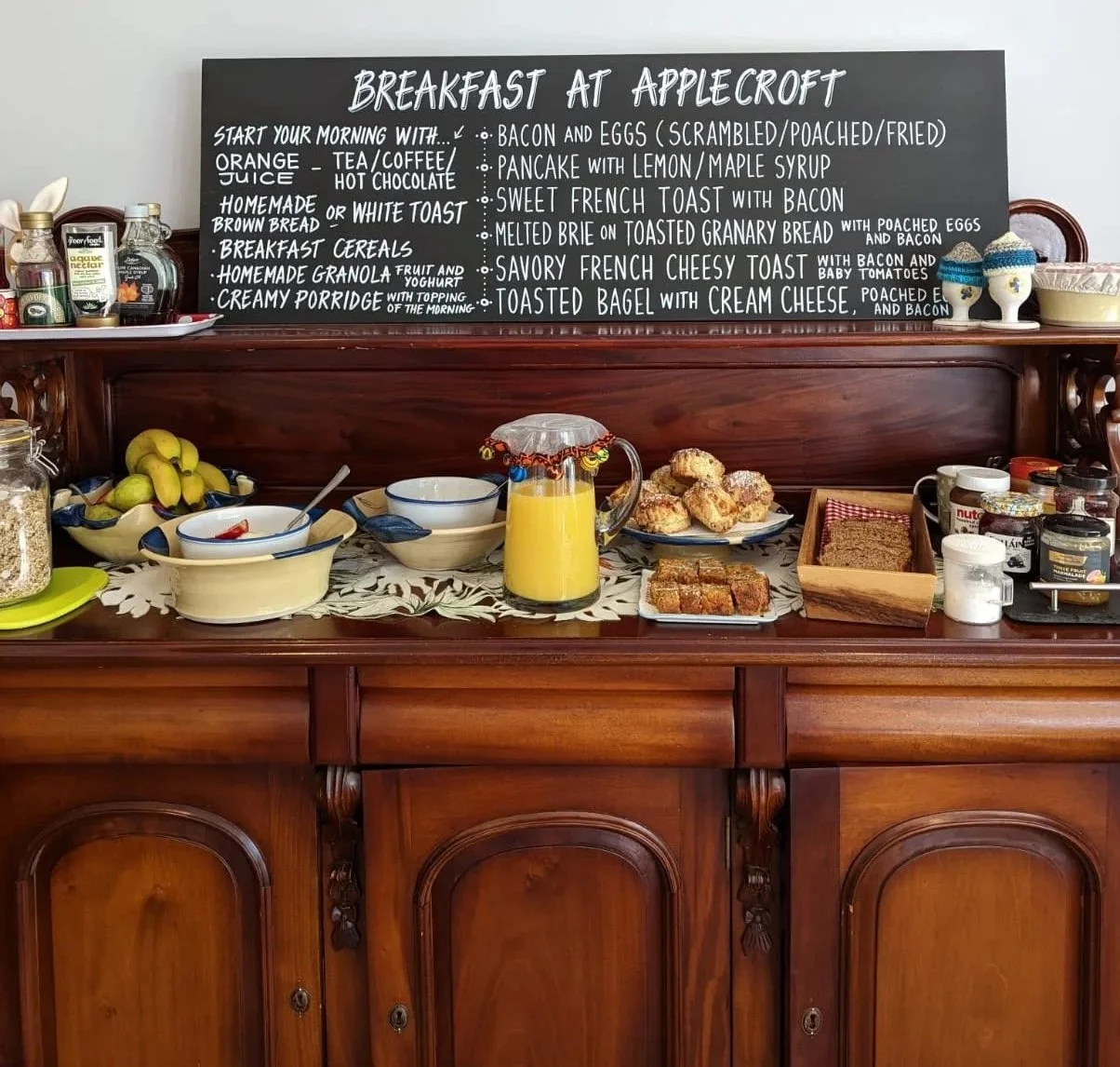 Breakfast spread on a wooden sideboard with a blackboard menu, bowls of cereal, a pitcher of orange juice, muffins, slices of bread, jars of spreads, and fresh fruit.