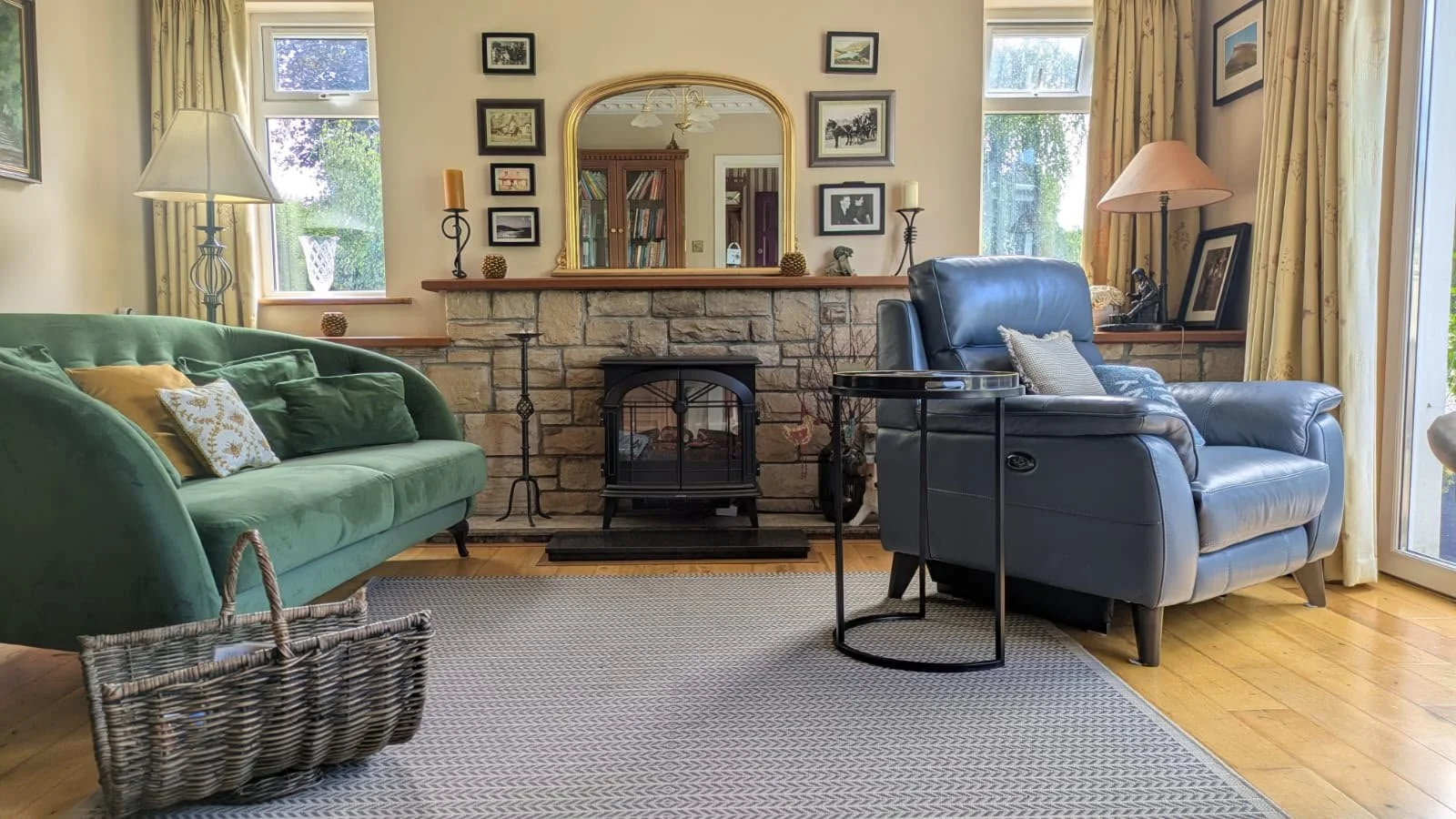 Living room with green sofa, black leather armchair, stone fireplace, mirror, framed artwork, and large windows with curtains, hardwood floors, and a patterned rug.