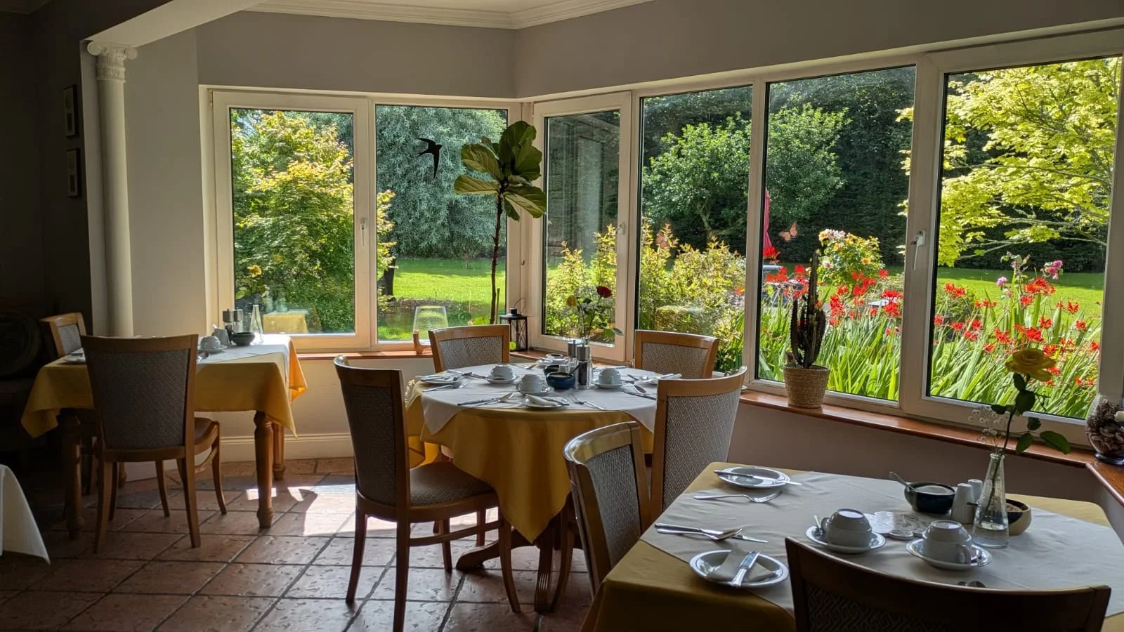 A cozy dining area with large windows showing a lush green garden outside. The tables are covered with yellow tablecloths, set with white dishes and silverware, and decorated with flowers in vases.