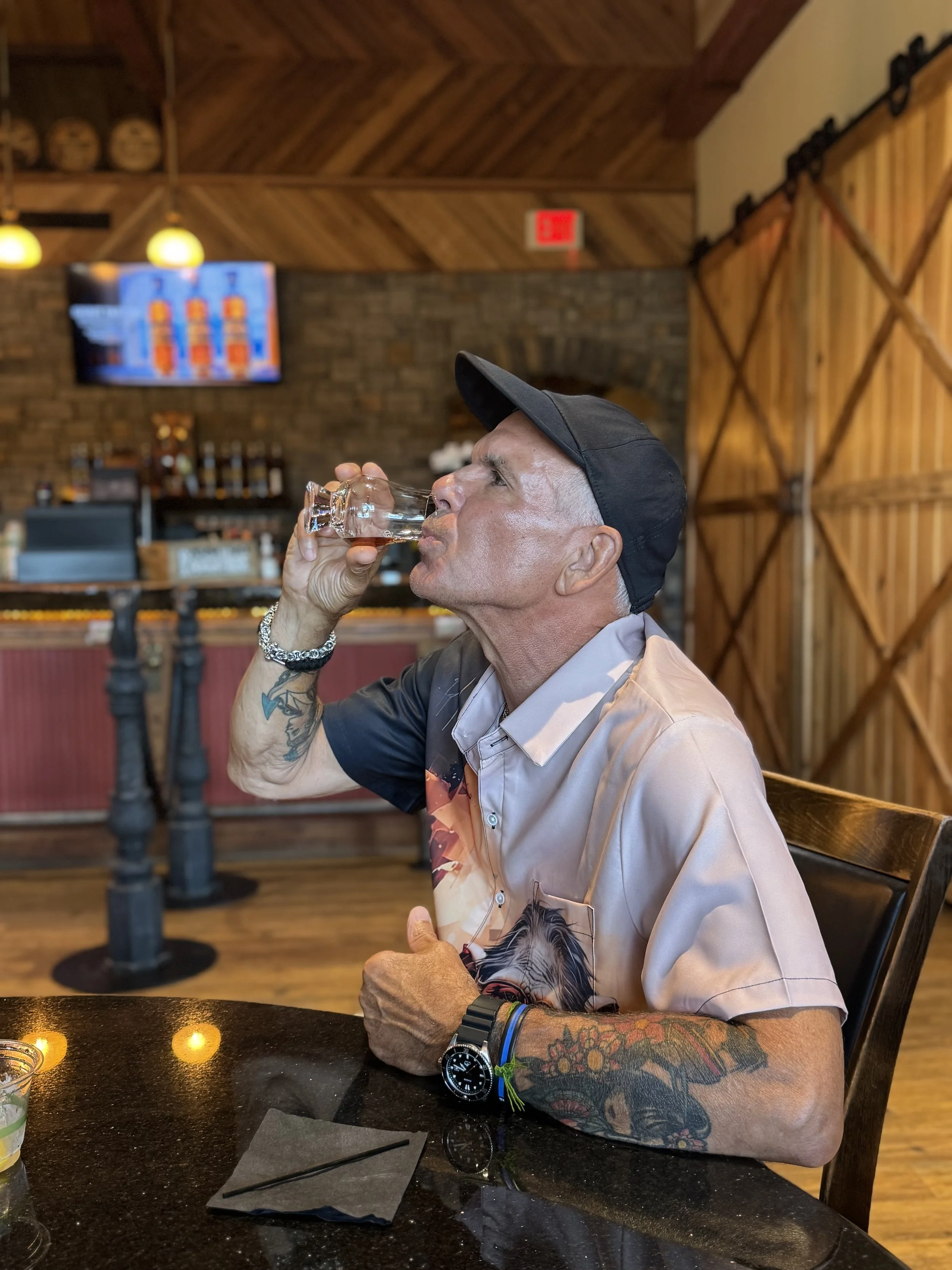 A man with tattoos on his arms and wearing a black cap is drinking from a shot glass in a rustic restaurant or bar setting. There are wooden walls and barrels visible. The table has glasses and a napkin with a straw.