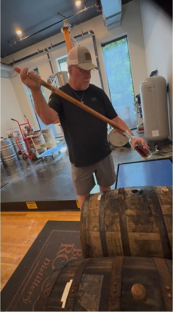 A man wearing a beige cap, black t-shirt, and shorts is pouring a barrel of whiskey through a long wooden tube into a glass, in a distillery or brewery setting.