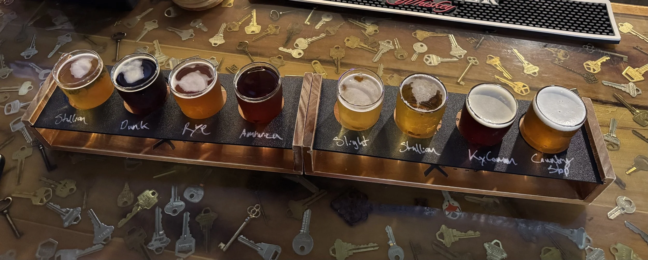 A flight flight tasting flight with eight small glasses of beer on a black tasting paddle, each labeled with a style or flavor like 'Shillian,' 'Dunk,' 'Rye,' 'Amber,' 'Light,' 'Shillian,' 'KyoGunn,' and 'Cinnamon.' The paddle is on a wooden table wi