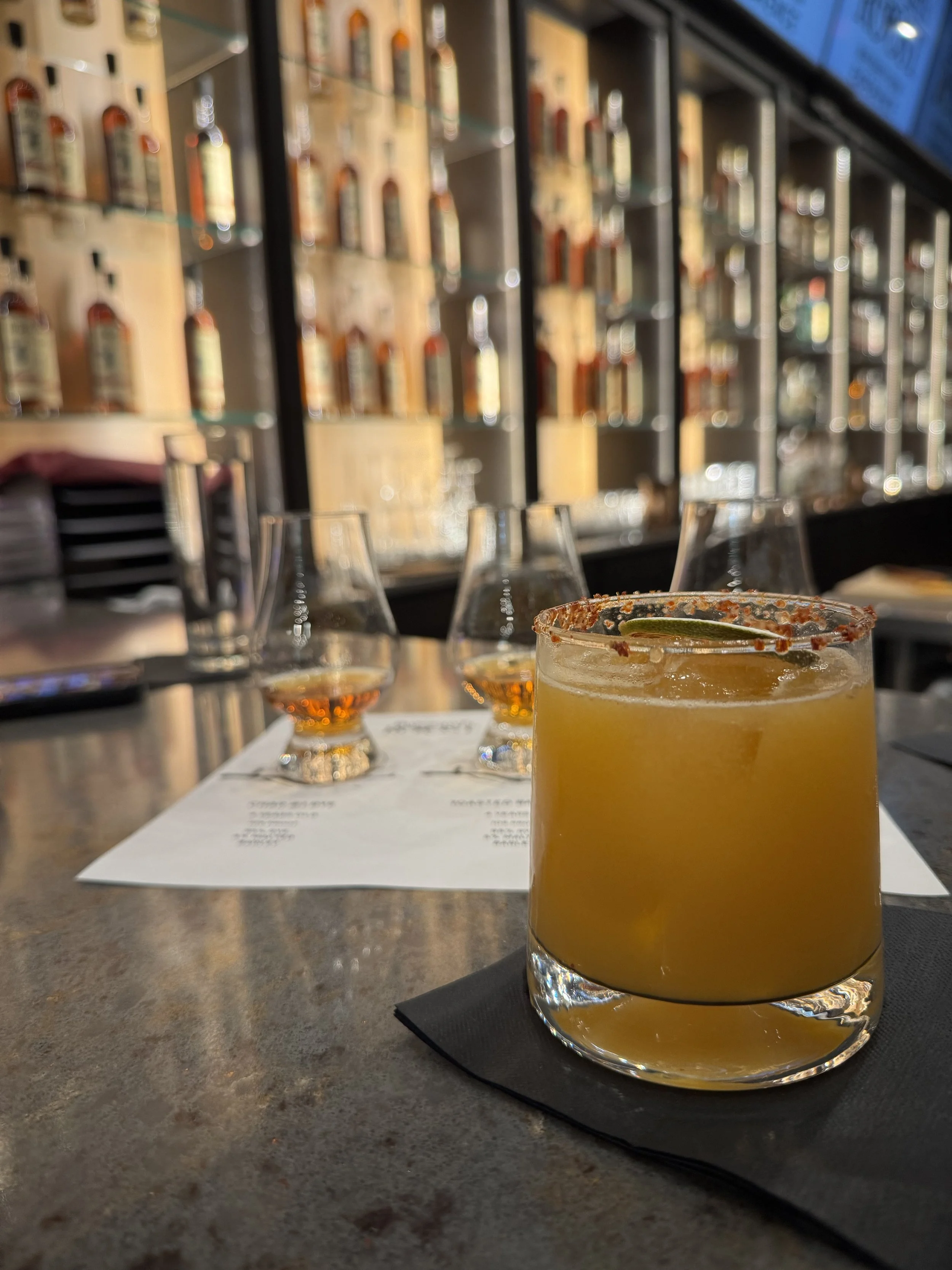 Close-up of a yellow cocktail with a salted rim and lime wedge on a black napkin, on a bar counter with glasses and a menu in the blurred background.