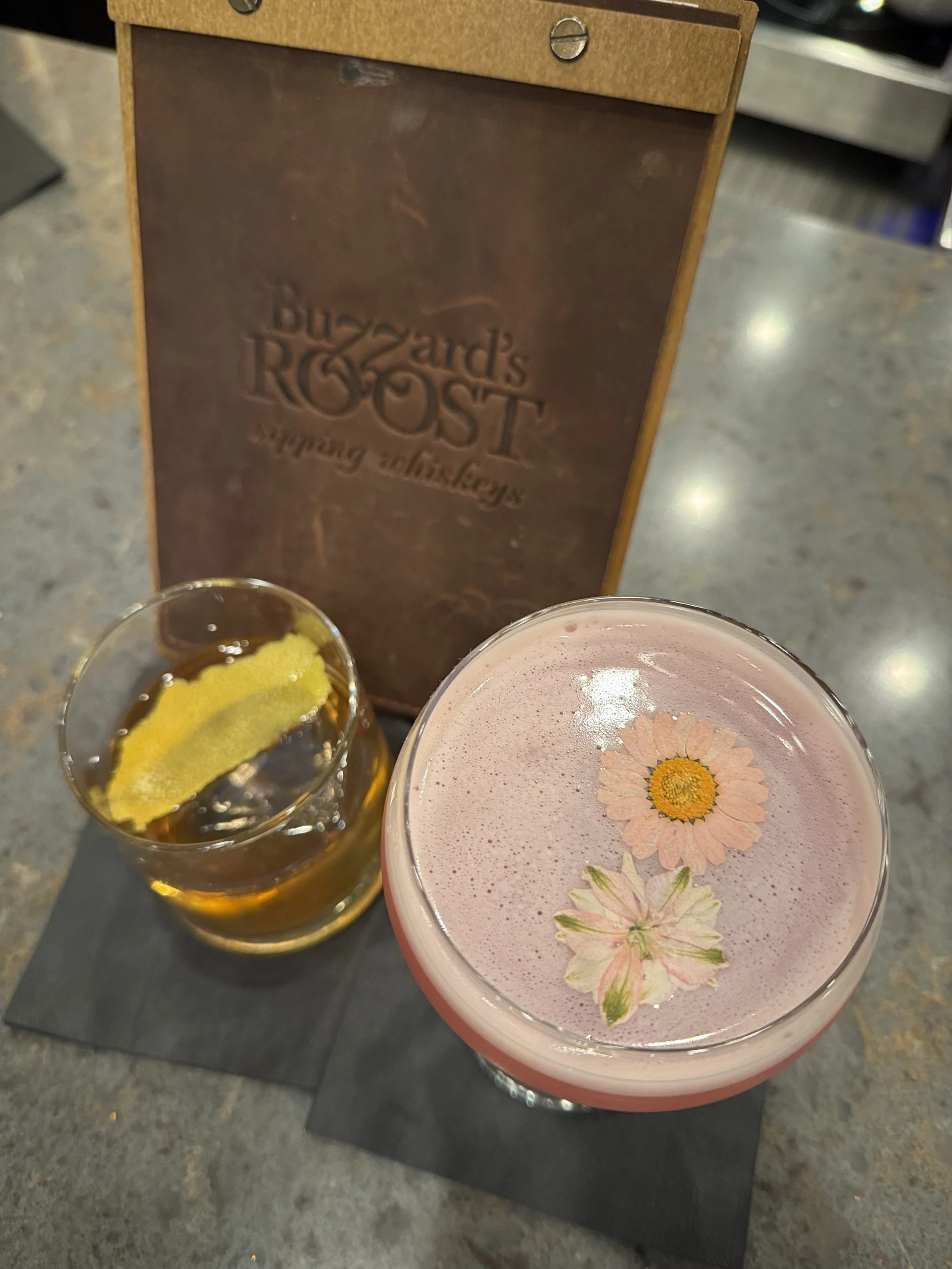A close-up of two drinks on a table, one is a glass of beer with lemon slices, and the other is a pink floral cocktail with a daisy flower decoration, with a Budweiser's ROST support sign in the background.
