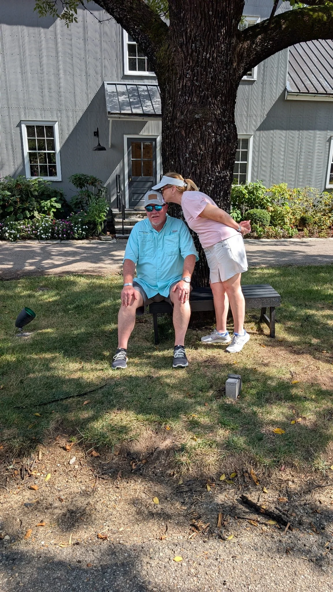 A man sitting on a black bench under a large tree, with a woman standing beside him, leaning in to give him a kiss on the forehead, in front of a gray house with white-framed windows and a garden.