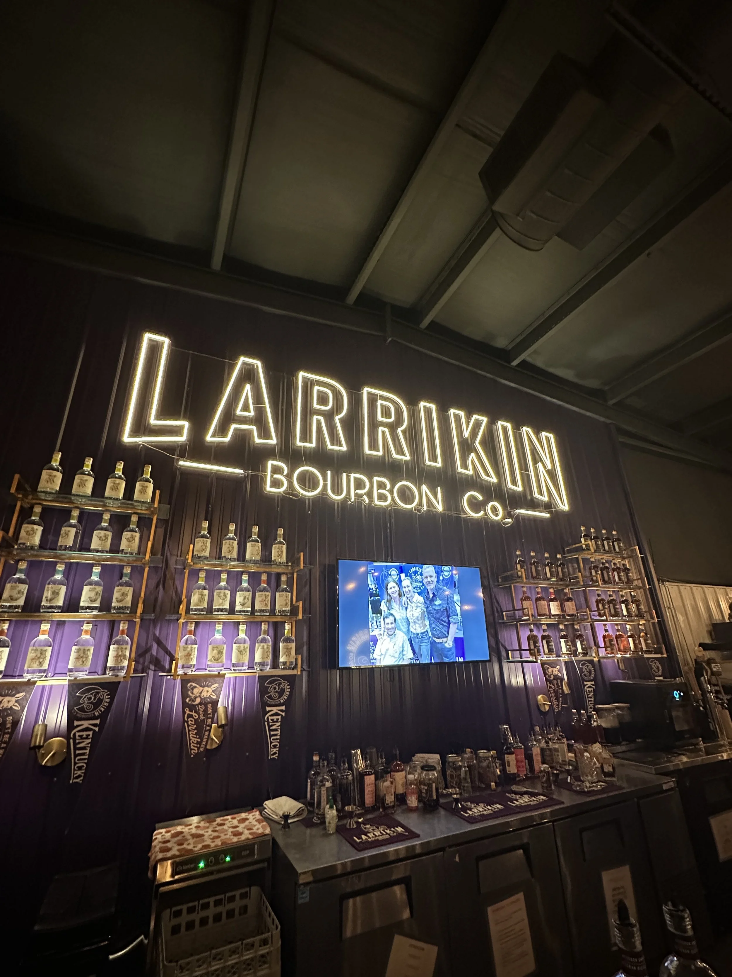 Impression of a bar with shelves of bourbon bottles, illuminated sign reading 'Larrikin Bourbon Co.', and a TV screen showing a group photo, with bar tools and glasses on the counter.
