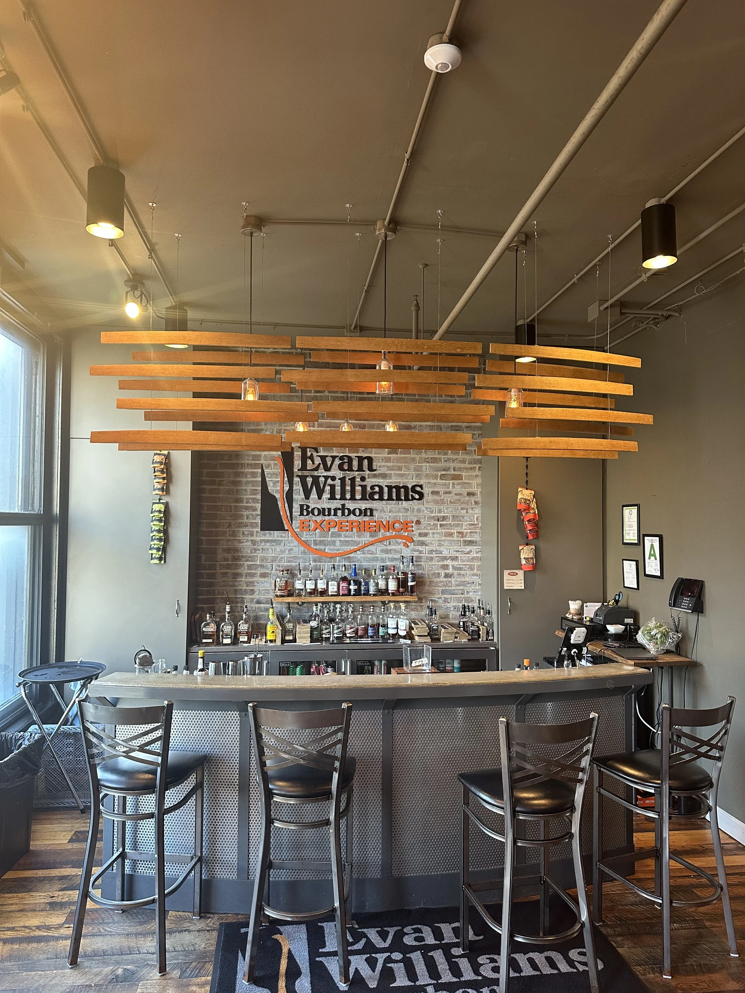Bar area inside Evan Williams Bourbon Experience with a brick wall, shelves of liquor bottles, a bar counter, and high chairs. Hanging wooden slats and pendant lights decoratively hang from the ceiling.