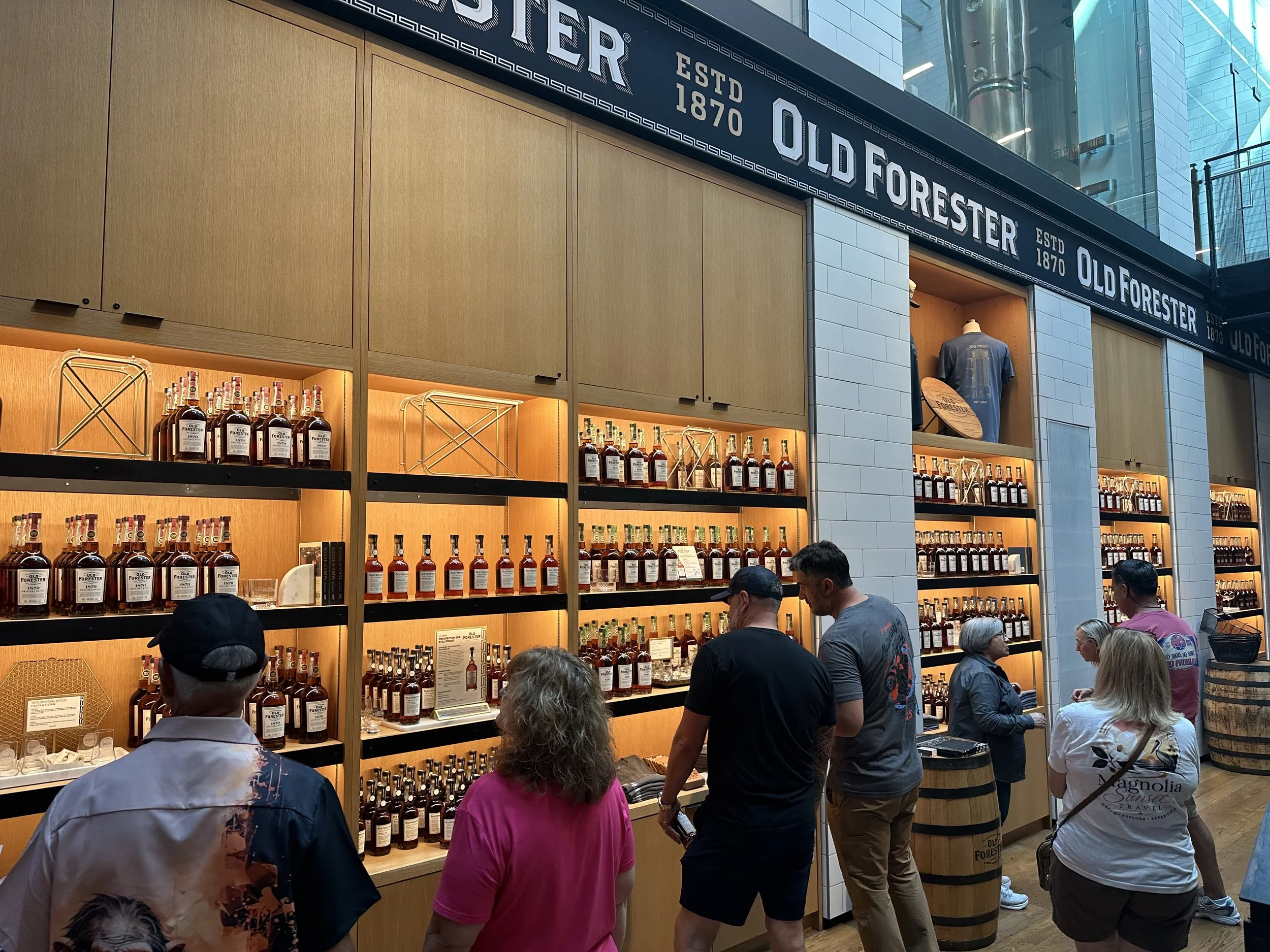 People shopping at a liquor store with wooden shelves filled with bottles of Old Forester bourbon whiskey, and a sign indicating the establishment was founded in 1870, inside a modern building with glass walls.