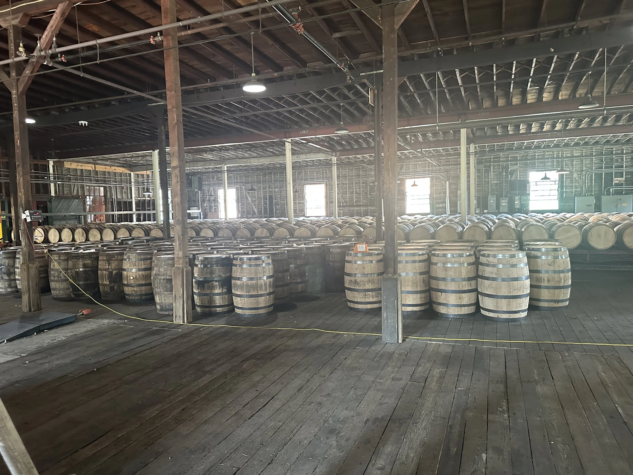A spacious warehouse with rows of wooden barrels, possibly for aging wine or spirits, and exposed wooden beams and metal piping on the ceiling.
