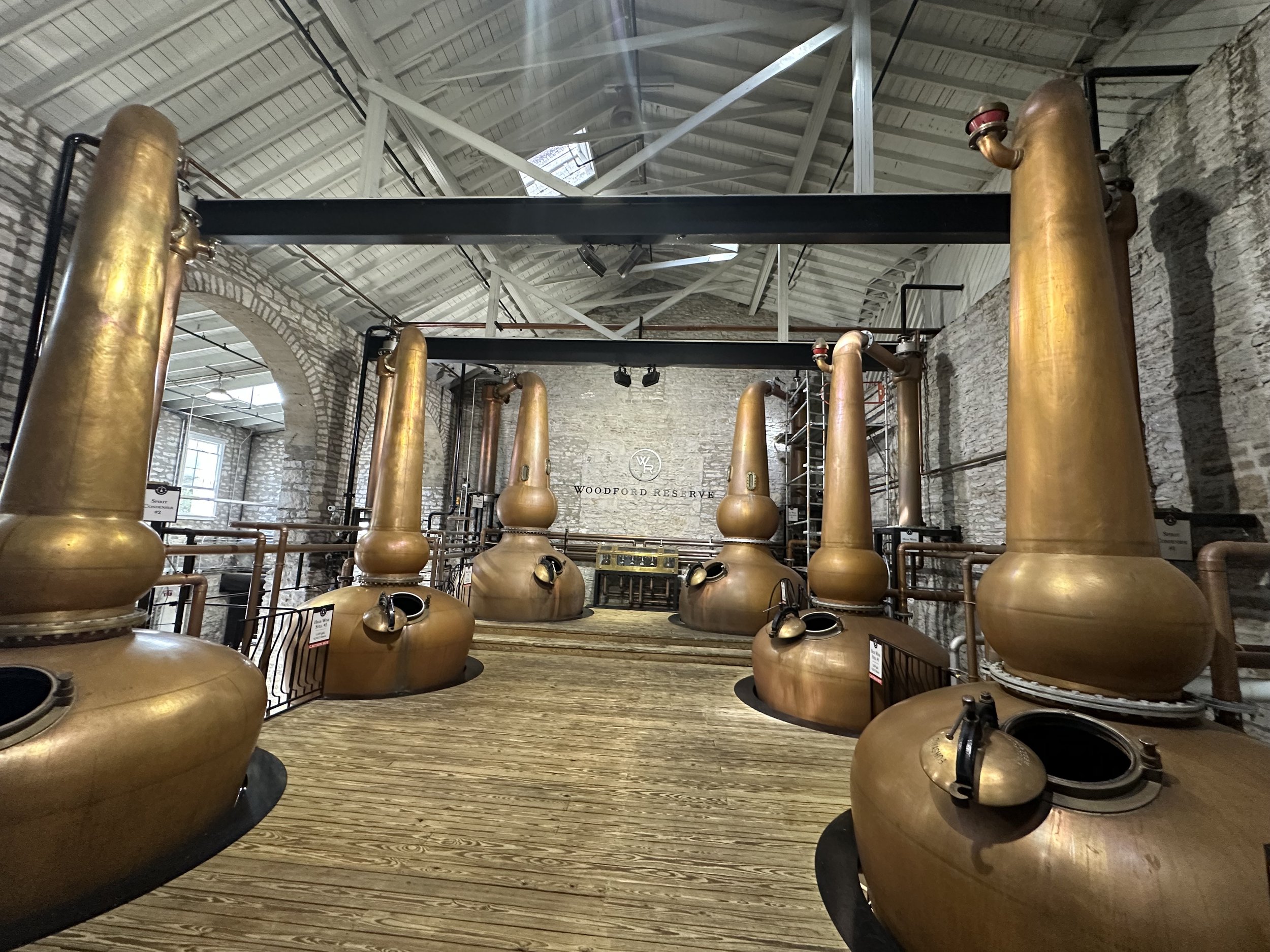 Interior of distillery with copper stills and stone walls, wooden floor, and high ceiling with metal beams.