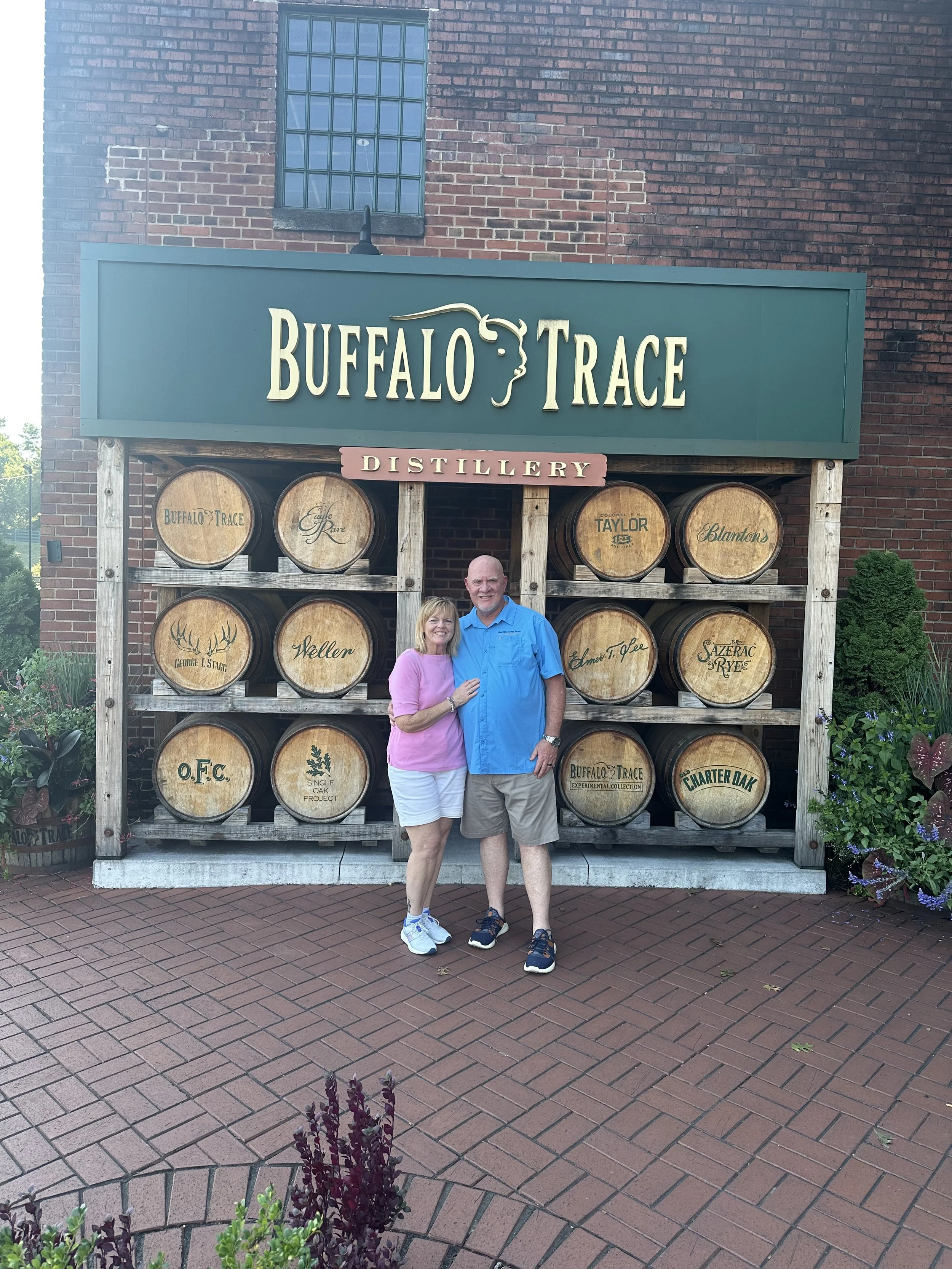 Two people, a woman and a man, standing together in front of a sign that reads 'Buffalo Trace Distillery'. They are smiling and have their arms around each other. The woman is wearing a pink shirt and white shorts, and the man is wearing a blue shirt