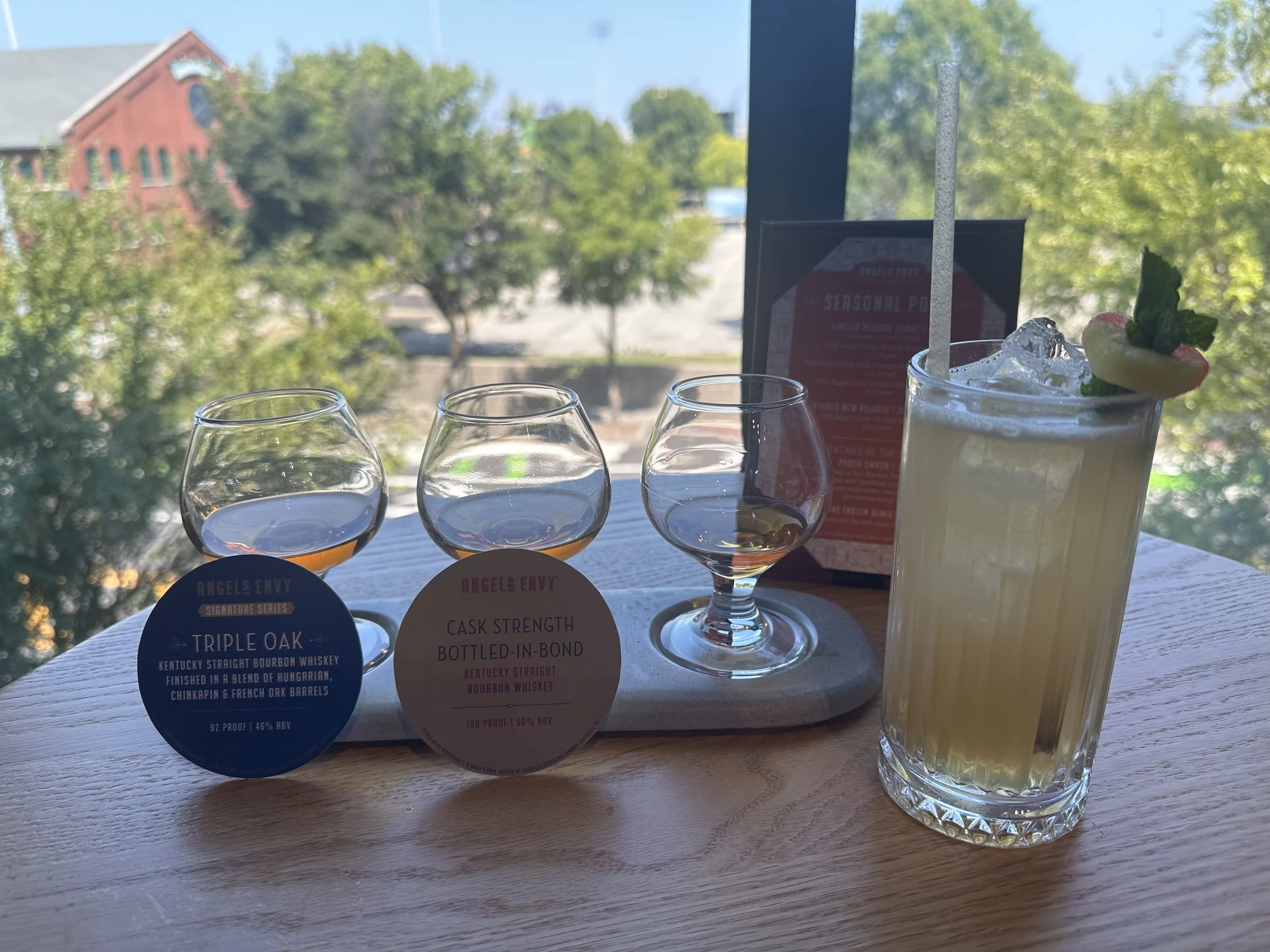 Three glasses of whiskey and a mixed drink on a wooden table with a view of trees and a building outside the window.