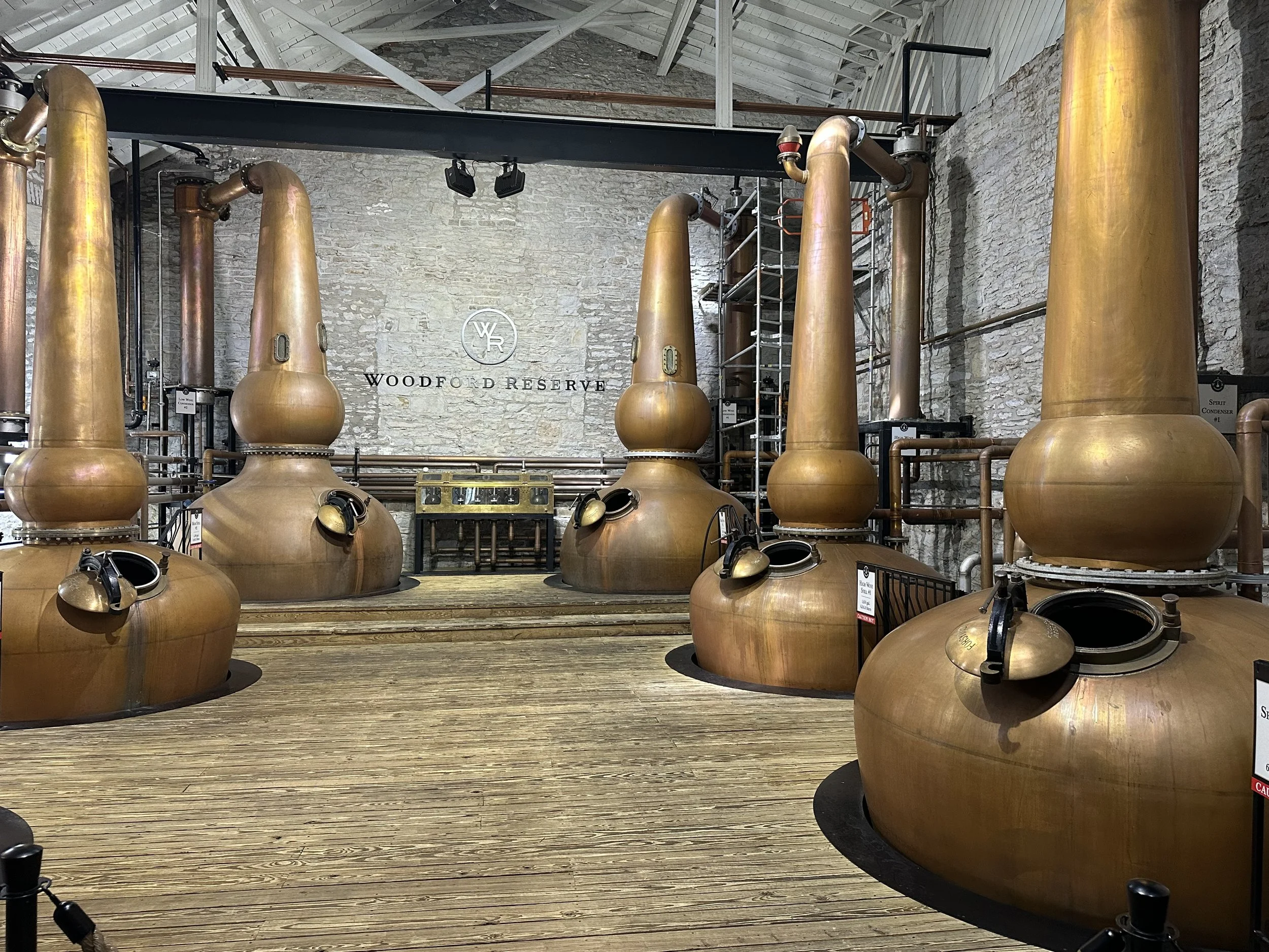 Distillery room with large copper stills, a stone wall, and a sign that reads 'Woodford Reserve'.