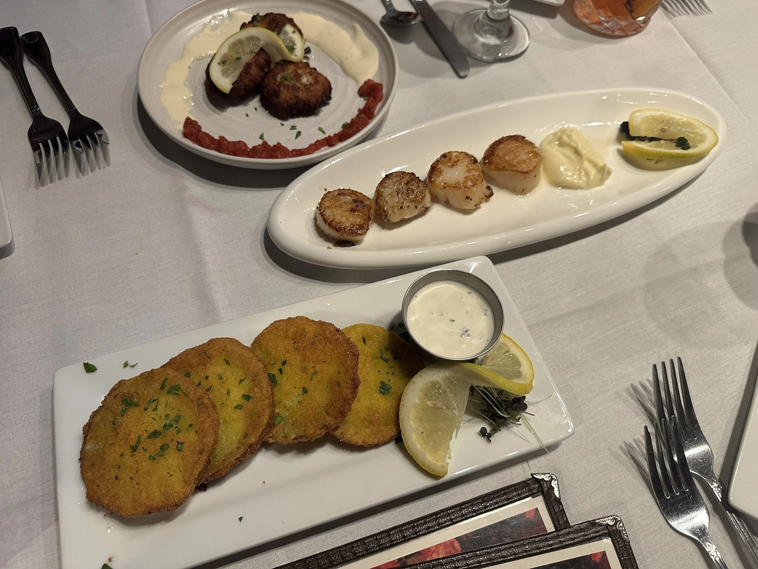 Fried scallops garnished with chopped herbs and lemon slices on a white rectangular plate, served with a side of tartar sauce. In the background, a plate with crab cakes topped with lemon wedges and sauce, and a small bowl of tartar sauce.