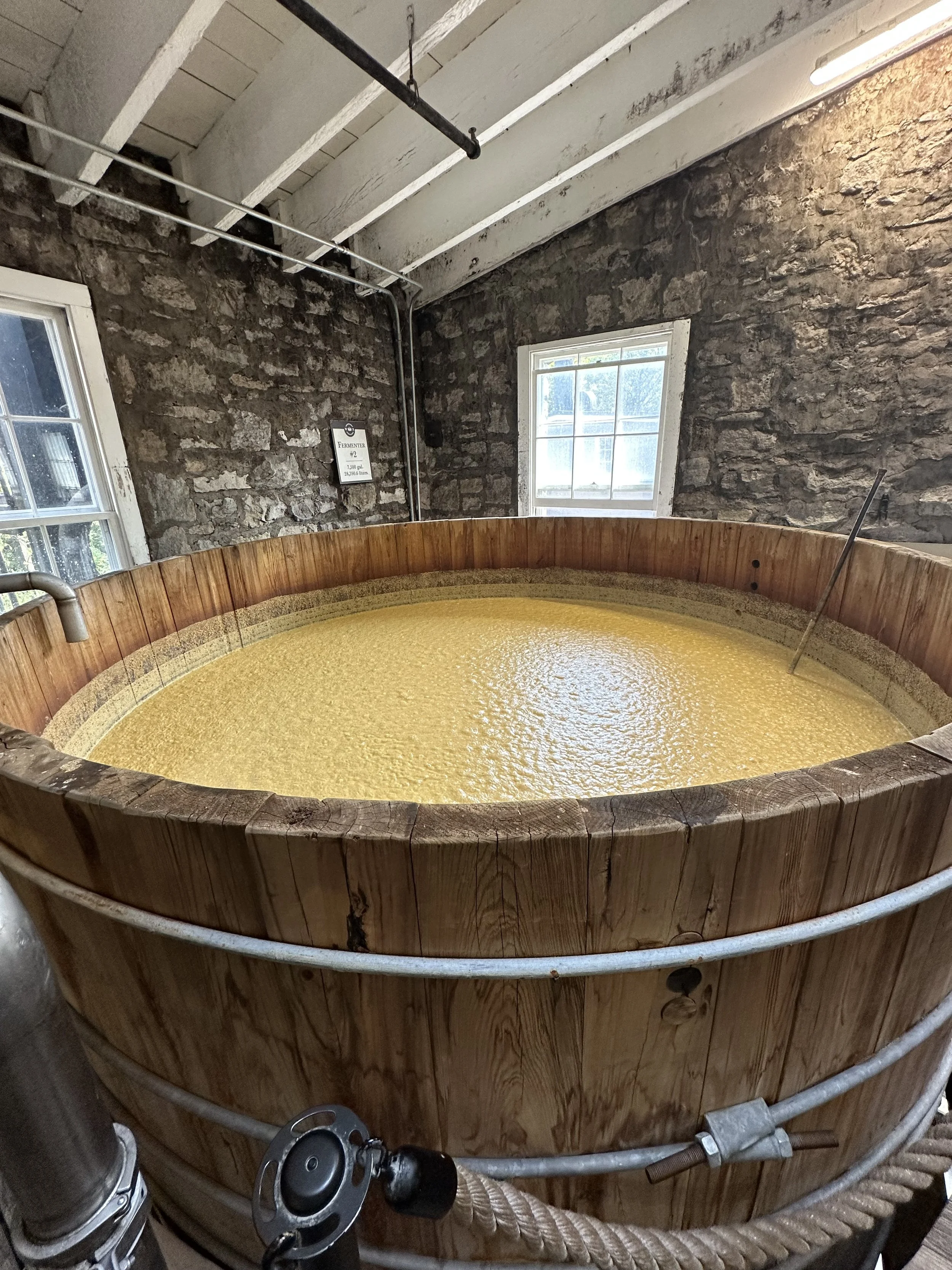 A large wooden tub filled with a yellowish liquid, situated in a room with stone walls, white ceiling beams, and a window. Pipes and metal fixtures are visible around the tub.