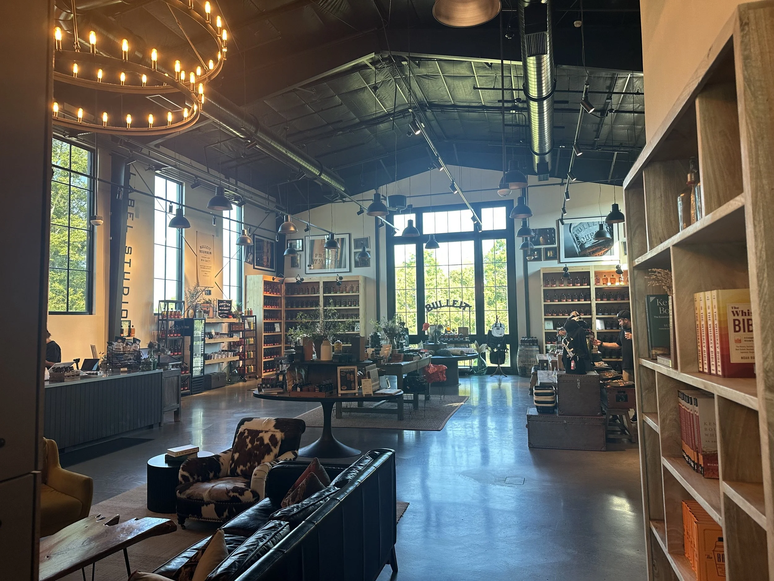 Interior of a modern retail store with large windows, black ceiling with exposed ducts and track lighting, wooden shelves filled with bottles and books, and seating area with a yellow chair and patterned armchair.