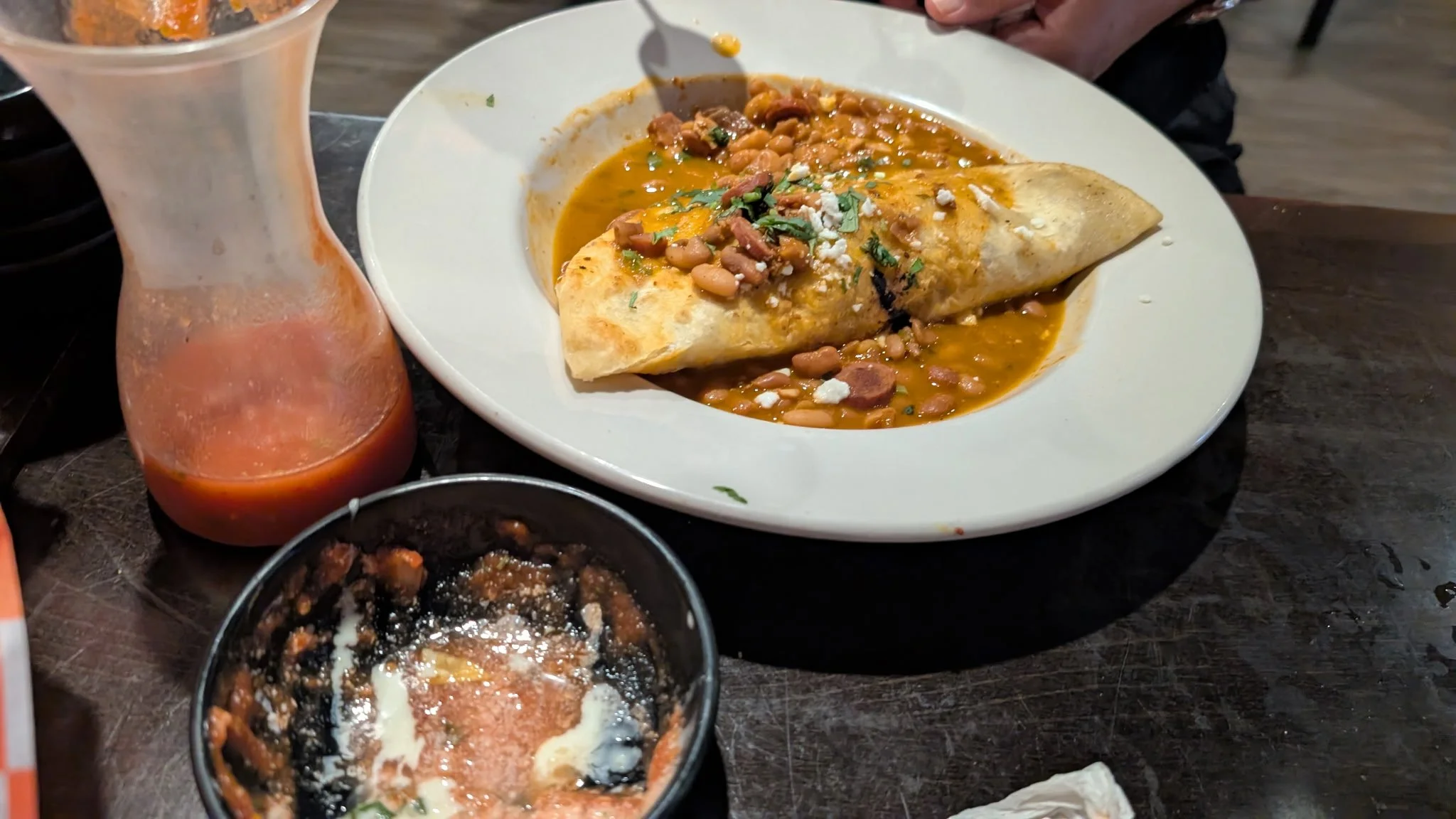 Plate of Mexican food with a burrito covered in beans and sauce, with a side of sauce and small bowls of beans, rice, and chili on a table.