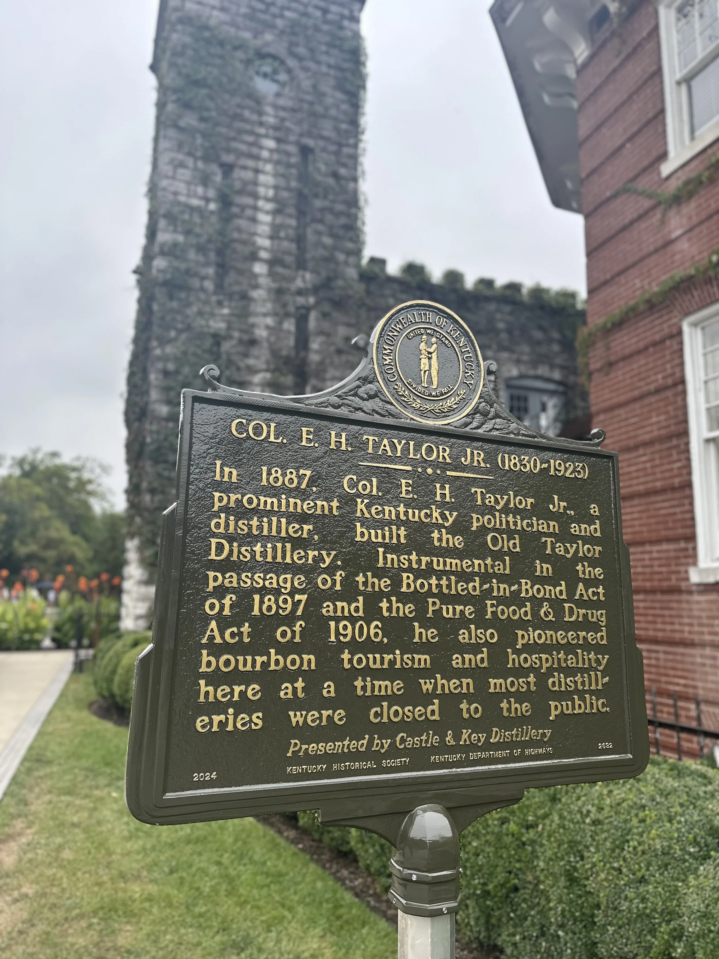 Historical marker about Col. E. H. Taylor Jr., a Kentucky politician and distiller, with a historic stone tower and brick building in the background.