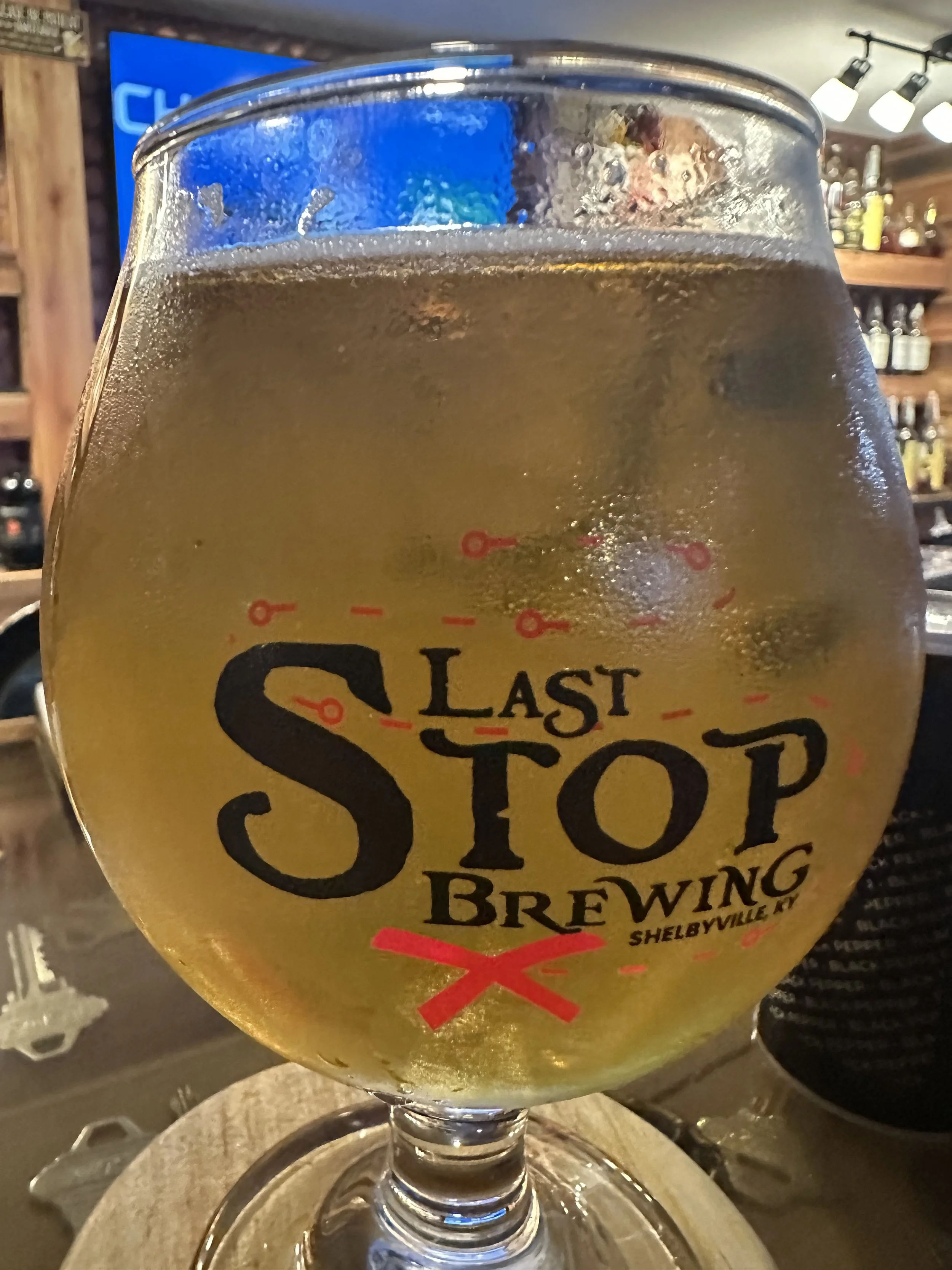 Close-up of a glass of cold beer with condensation on the surface, with a label reading 'Last Stop Brewing' and a bar setting in the background.