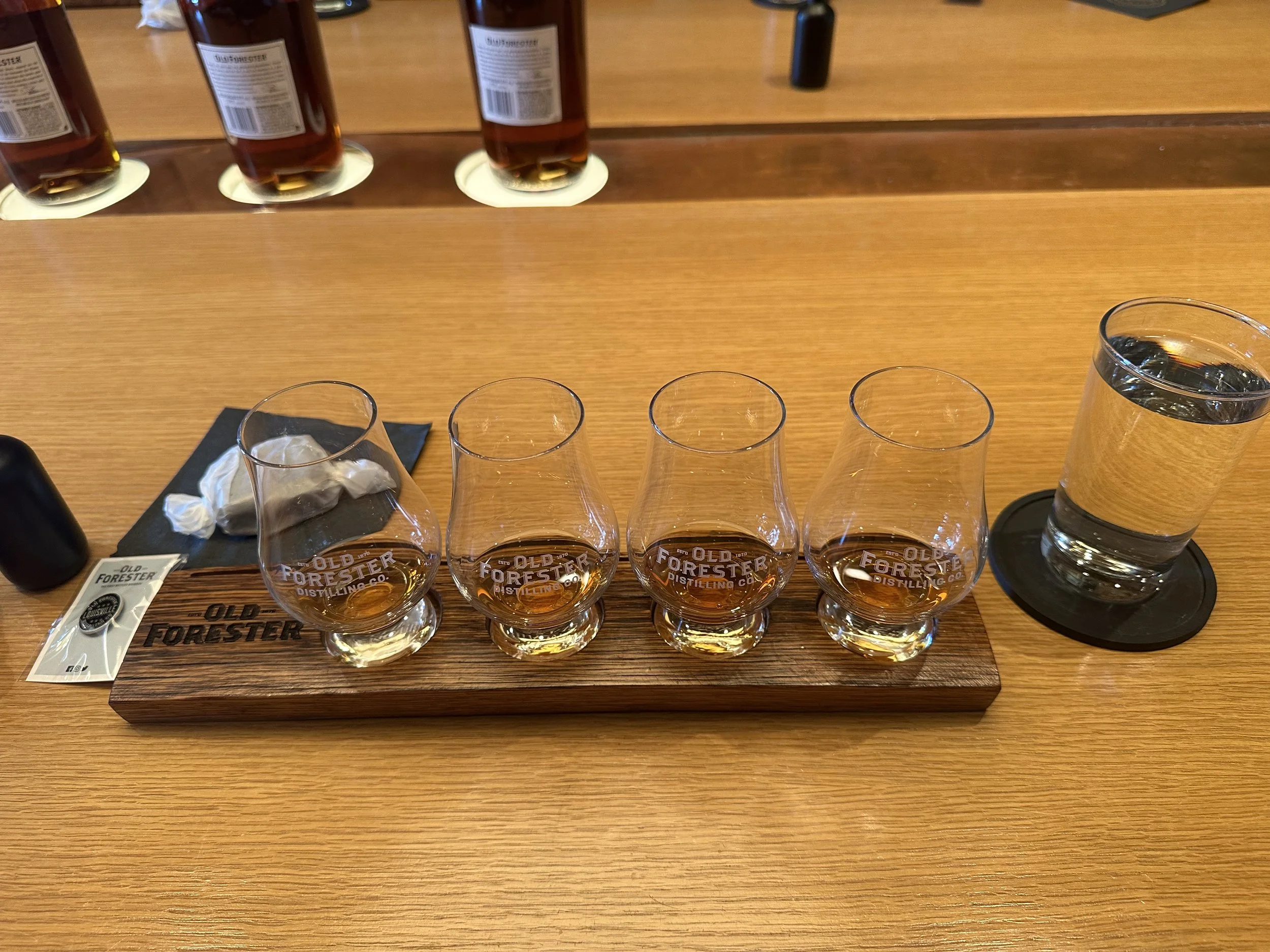 Four Glencairn glasses filled with whiskey on a wooden tray with 'Old Forester' branding, placed on a wooden table. Four bottles of whiskey are visible on a shelf in the background.