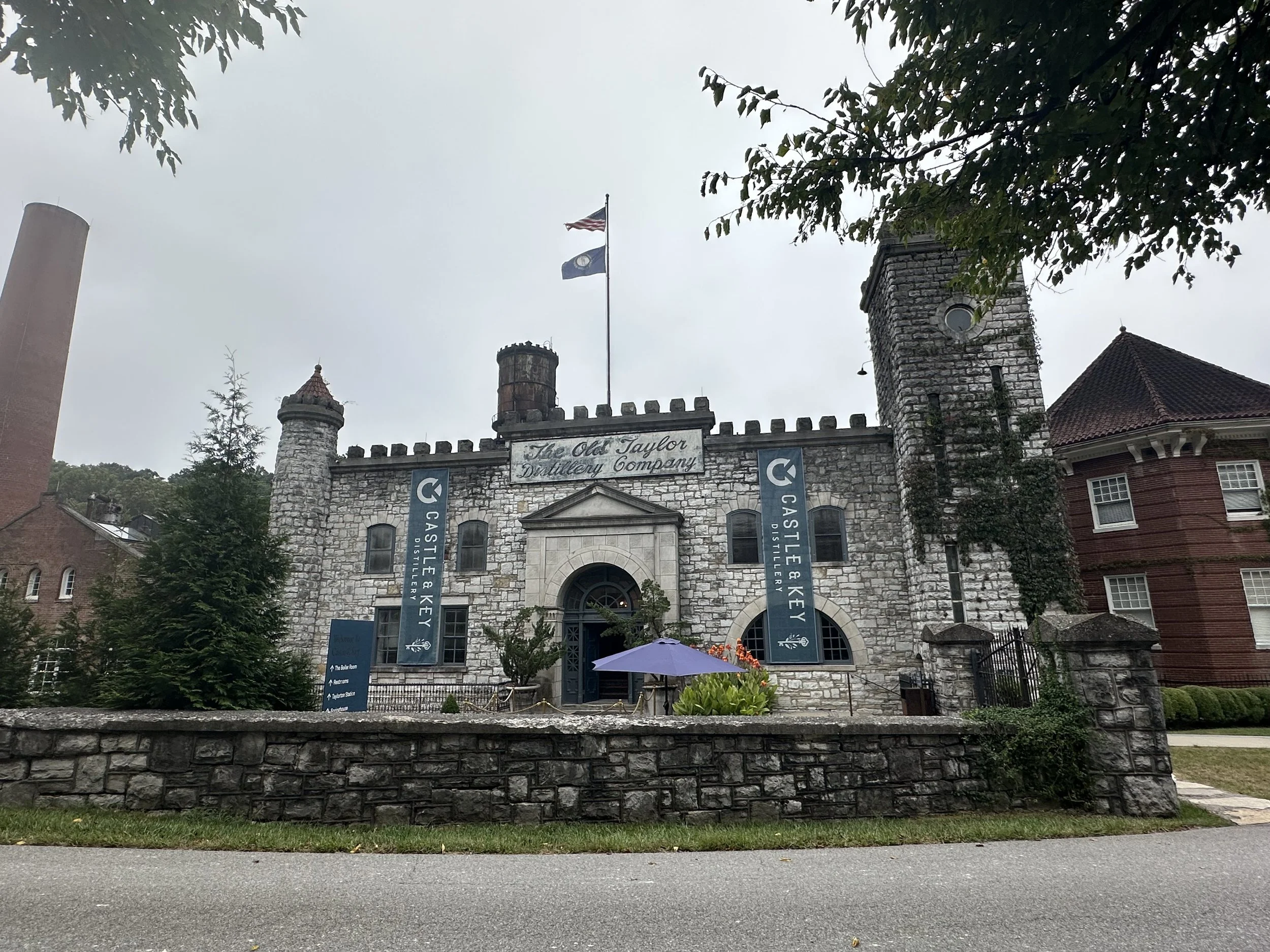 The Old Taylor Distillery Company building, made of stone with two turret towers, has flags flying on top and banners hanging on the front. There are trees and a sidewalk in front of the building.