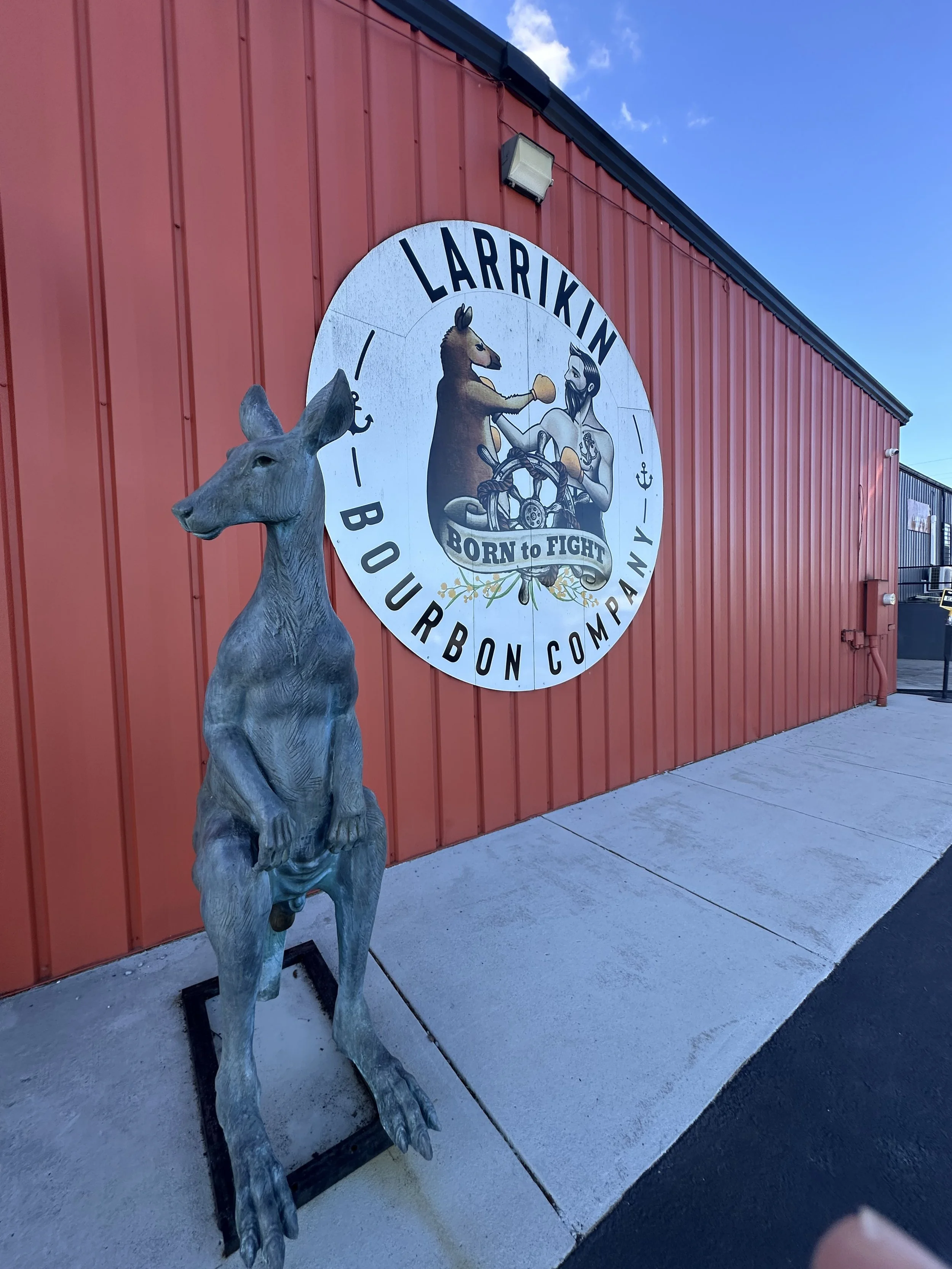 A life-sized sculpture of a deer placed outdoors near a red metal building with a large circular sign featuring a cartoon-style image of a horse and a man in boxing gloves, with the text 'LARRIKIN BOURBON COMPANY' and a banner saying 'BORN TO FIGHT'.