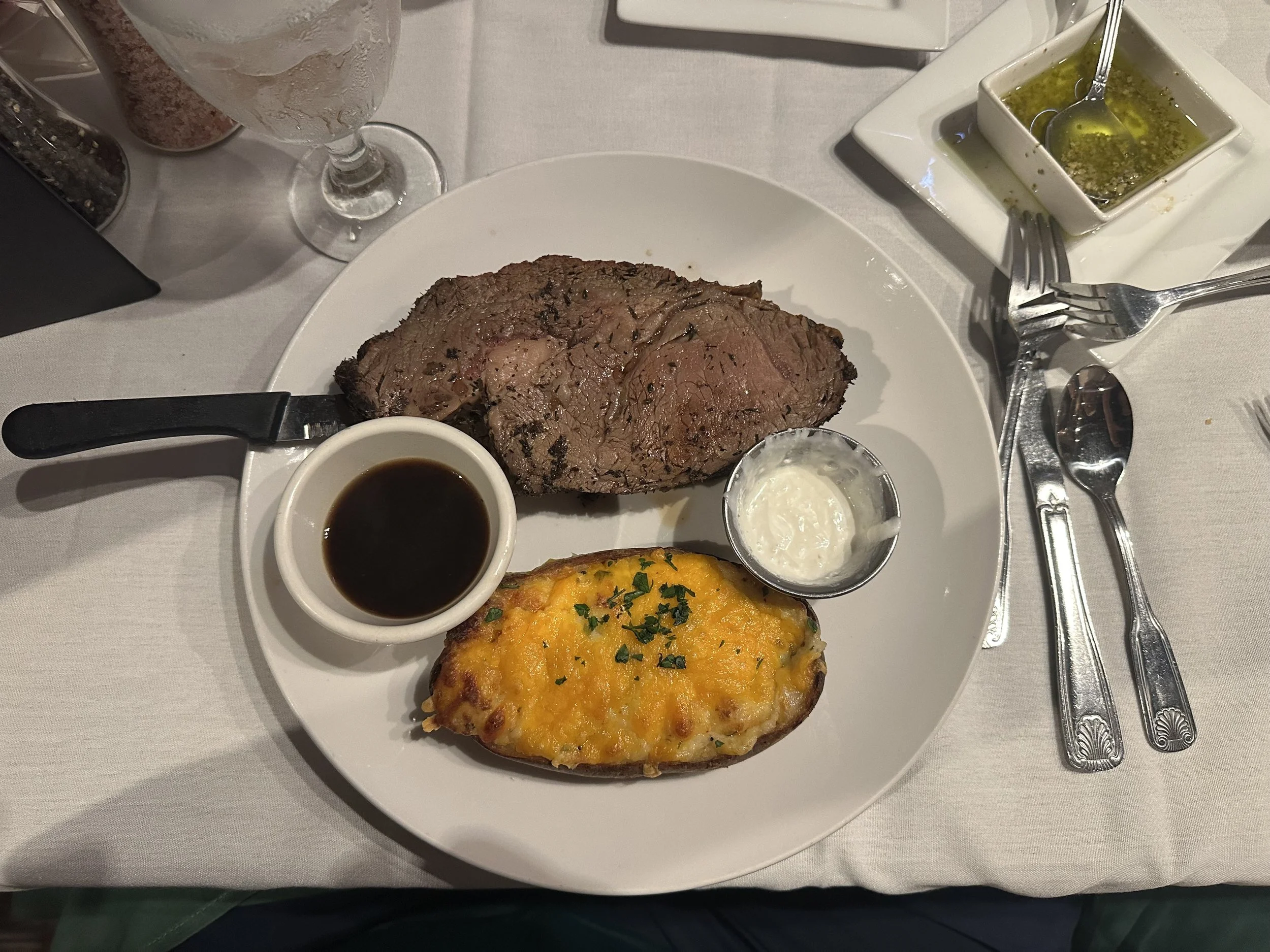 A plate with a cooked beef steak, a baked potato topped with cheese and herbs, and small bowls of sauce, with a glass of water, a small bowl of green herb oil, and condiments on the table.