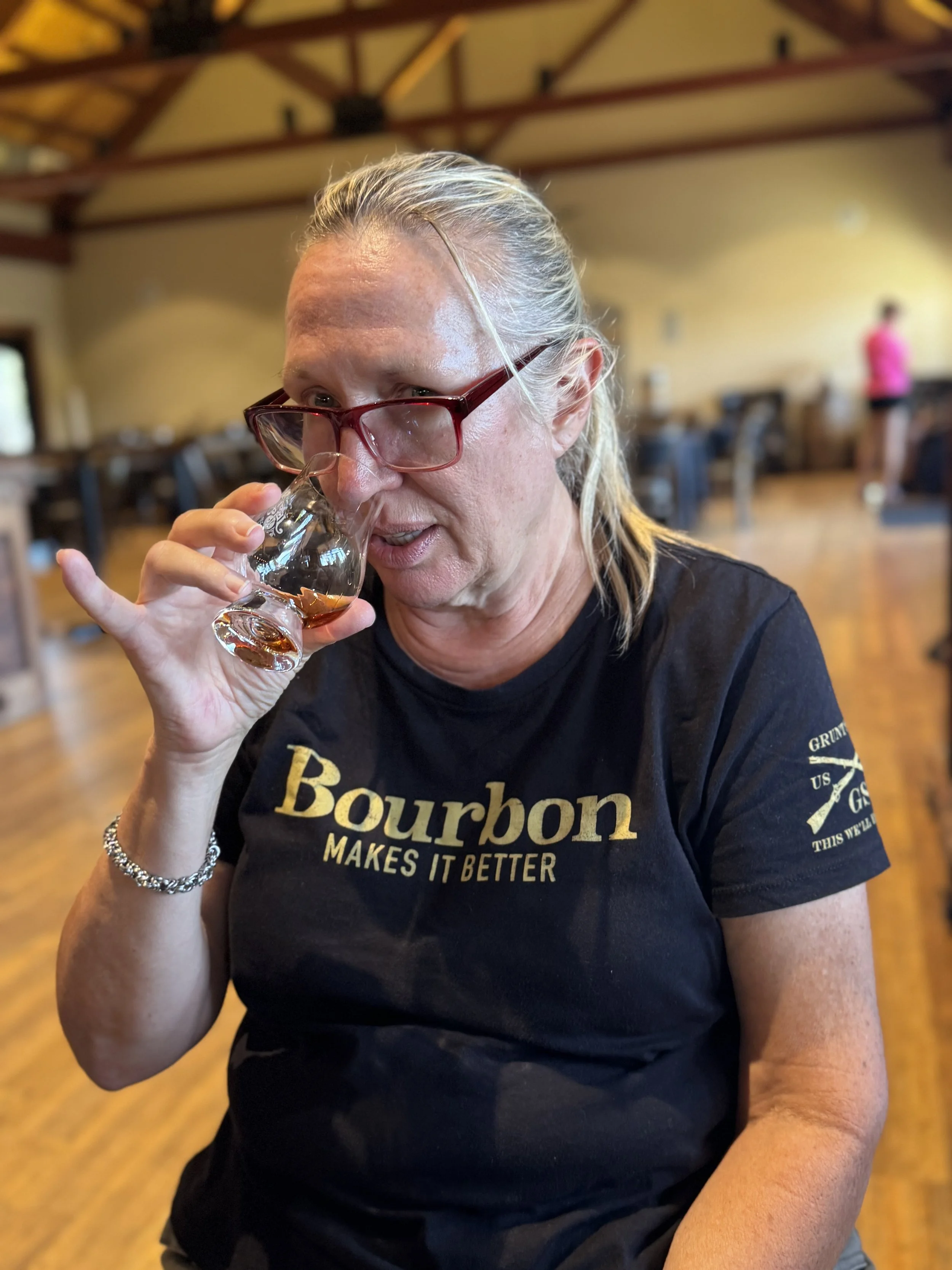 A woman with gray hair, glasses, and a black Bourbon shirt is drinking from a glass of whiskey or bourbon in a restaurant with a wooden floor and yellow walls.