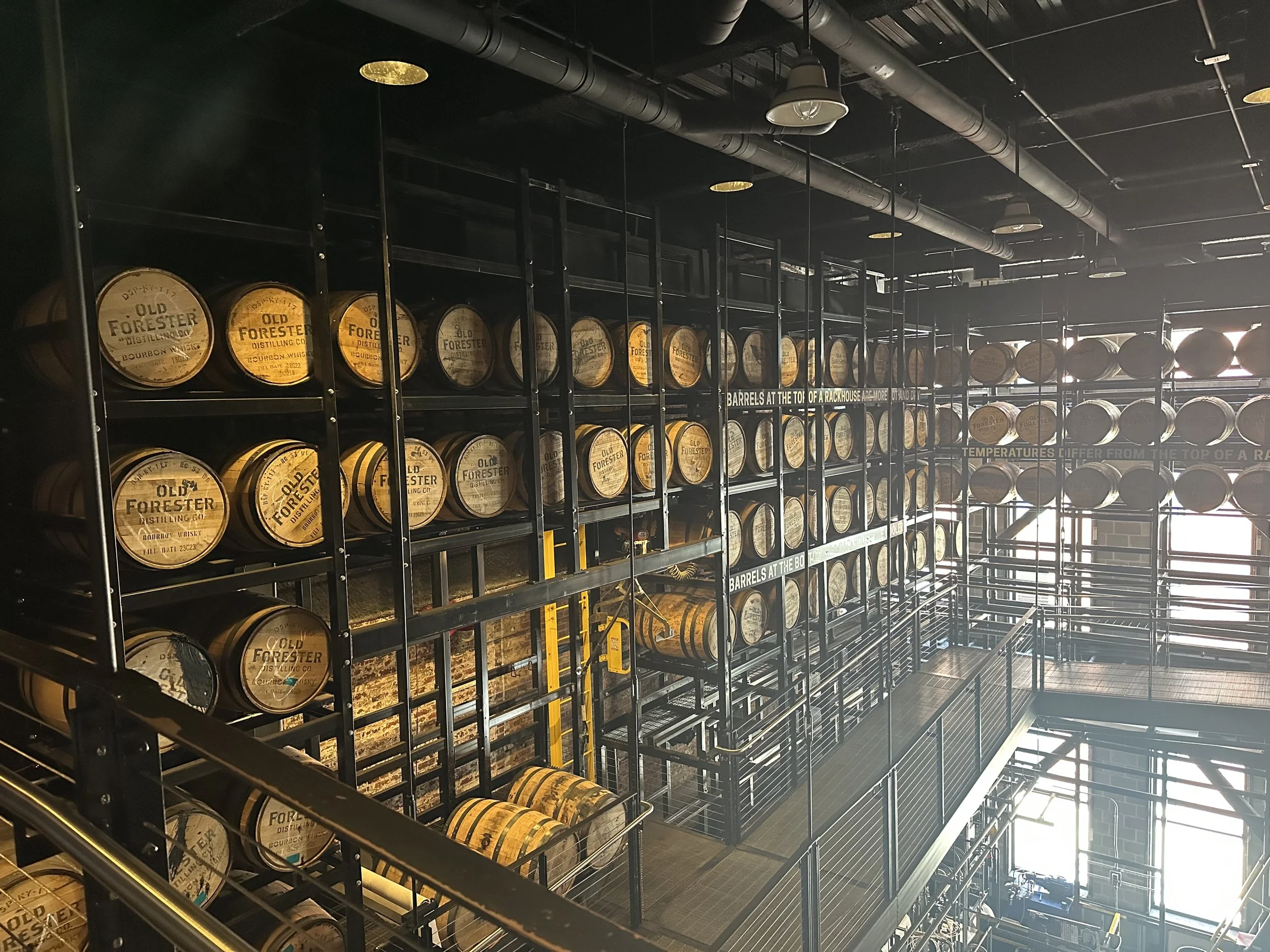 Multiple barrels of bourbon whiskey stacked on metal shelves inside a distillery, with industrial pipes and lighting overhead.