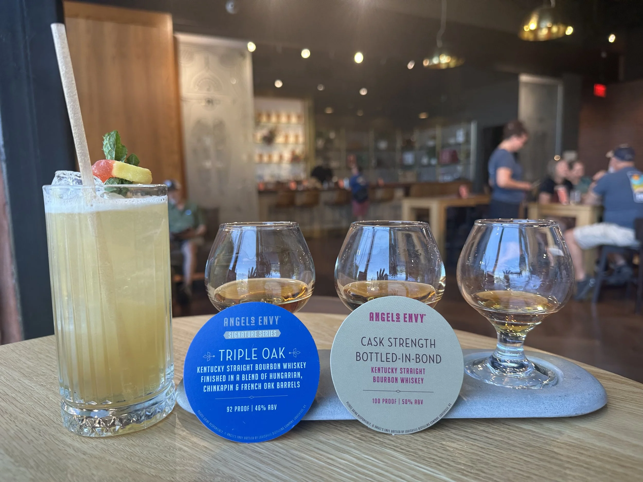 Focus on a tall glass of yellow cocktail garnished with mint and fruit, next to three whiskey glasses, and informational tags about Kentucky straight bourbon whiskey, on a wooden table inside a bar or restaurant with blurred background of people dini