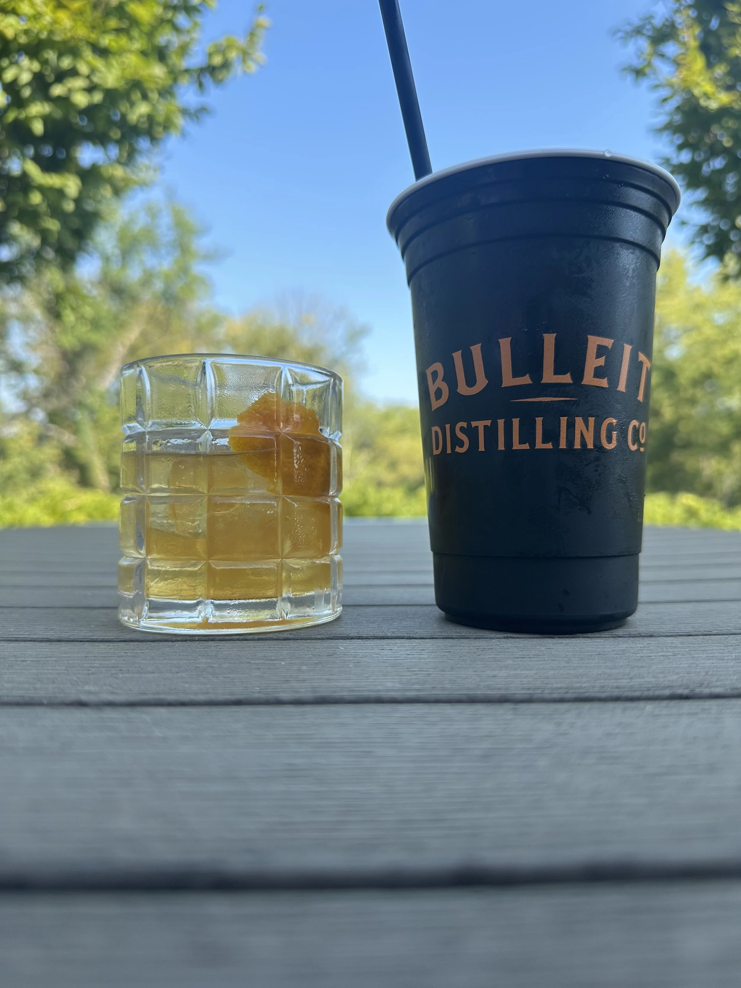 A black plastic cup with a straw and a clear glass filled with a yellow beverage and orange slices, set on a light-colored wooden surface outdoors with a background of green trees and a blue sky.