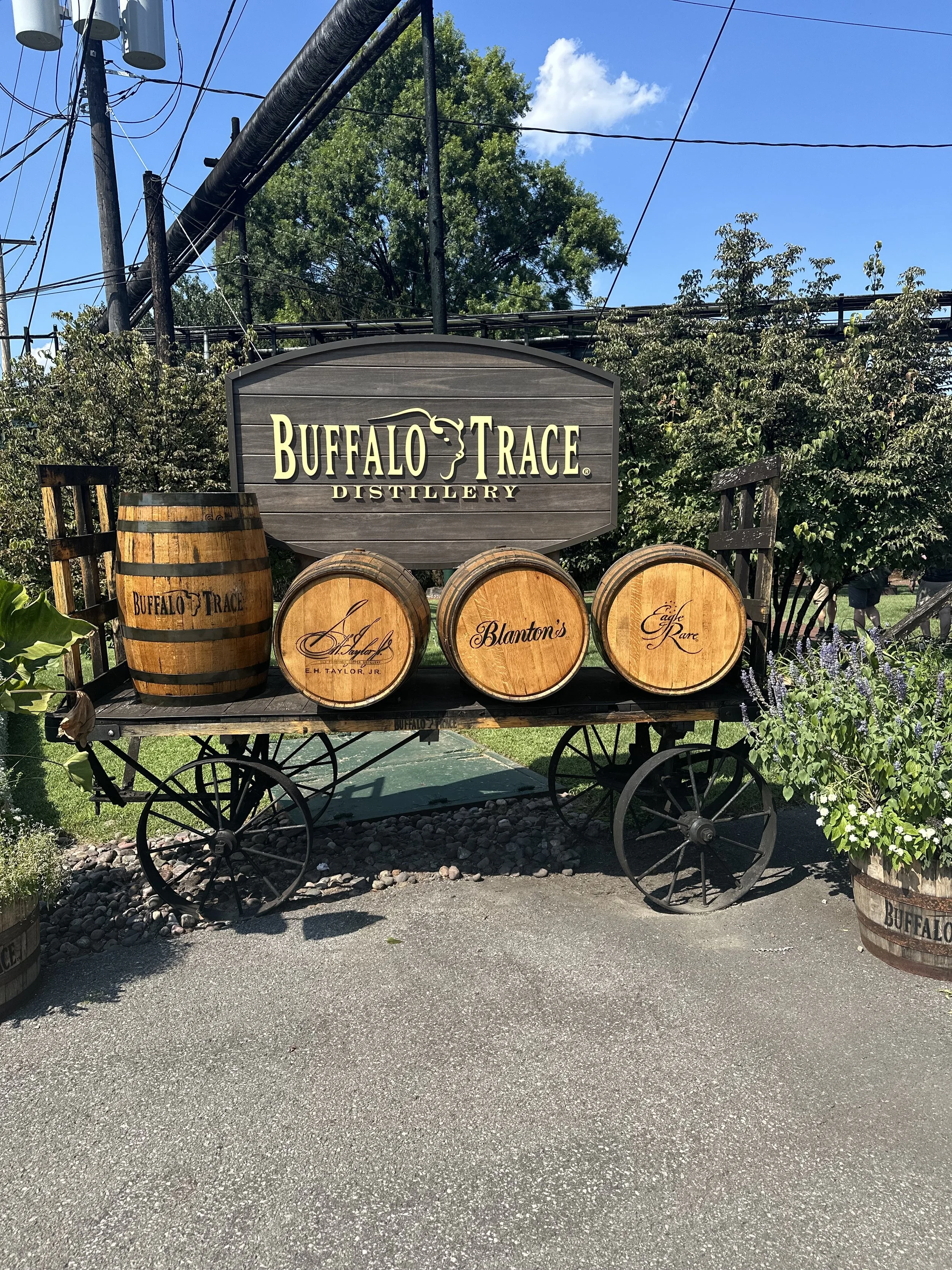 Wooden cart with barrels and a sign for Buffalo Trace Distillery, surrounded by greenery, under a blue sky with clouds.