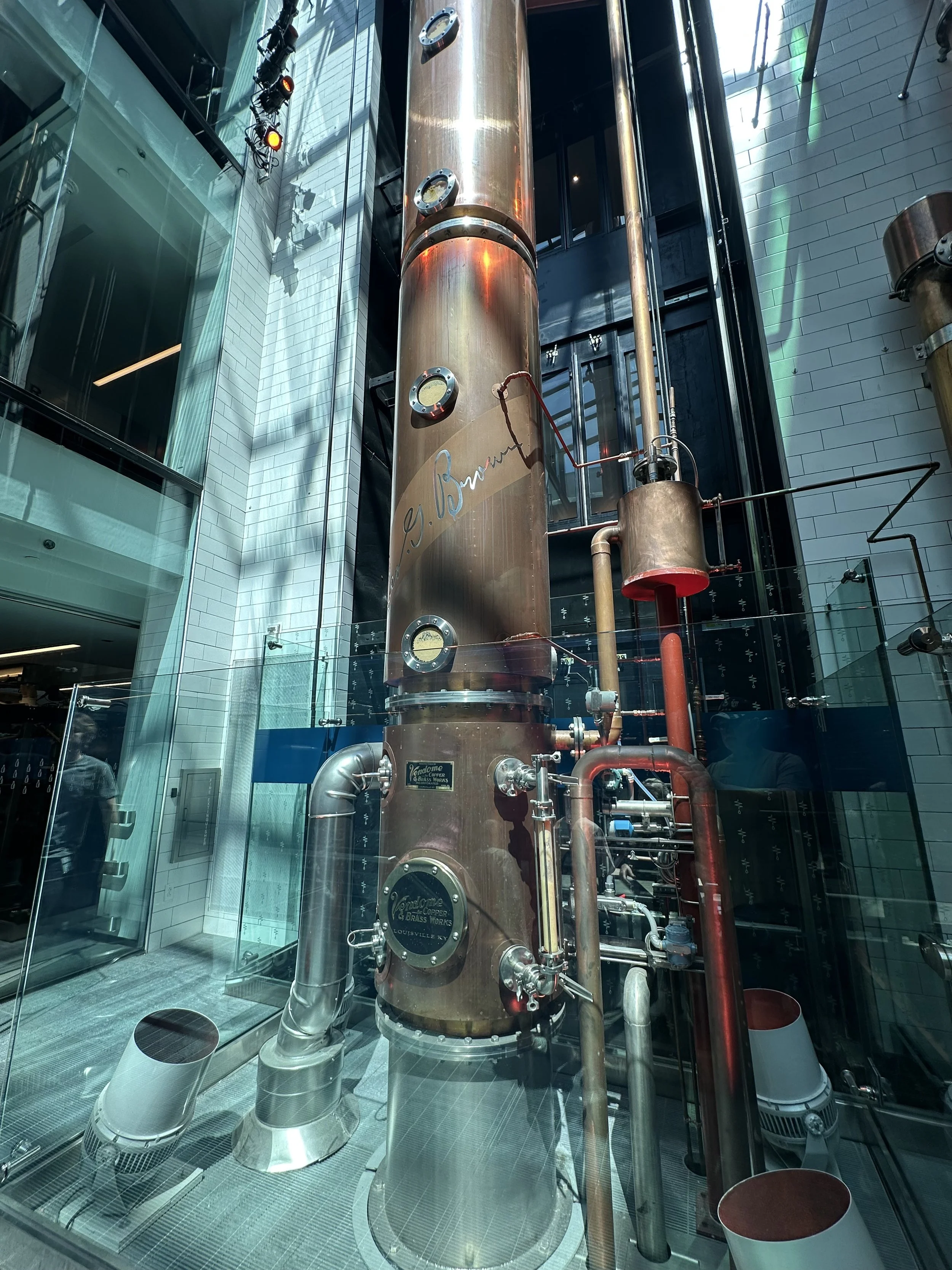 A large, vertical copper distillation column inside a modern building with glass walls, pipes, gauges, and industrial equipment, displayed behind a glass barrier.