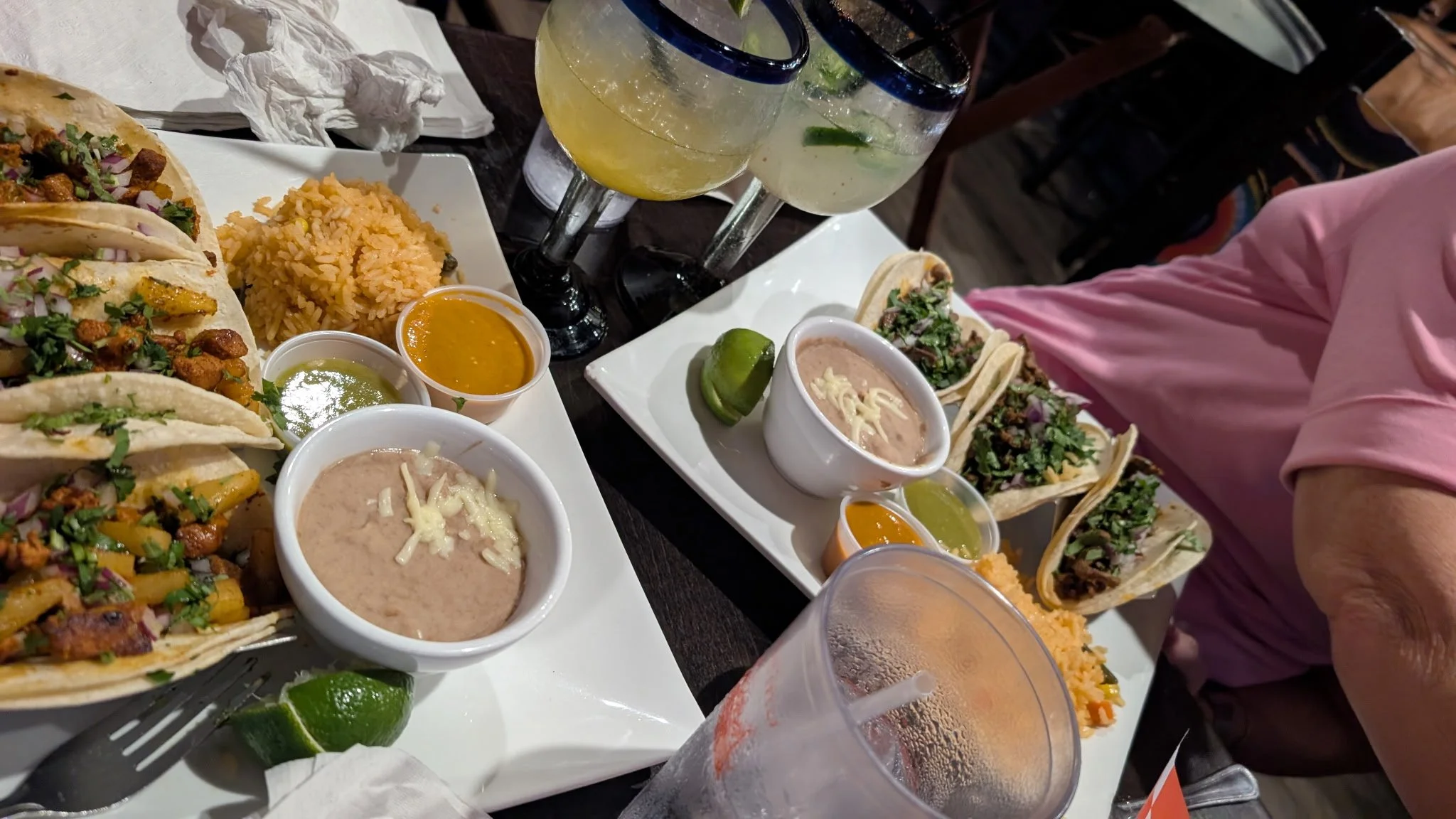 Three plates of tacos with various fillings, topped with cilantro and onions, some with shredded cheese and meat. Small bowls of condiments such as green sauce, orange sauce, and sour cream. A plate of rice. Two margarita glasses with lime slices. A 