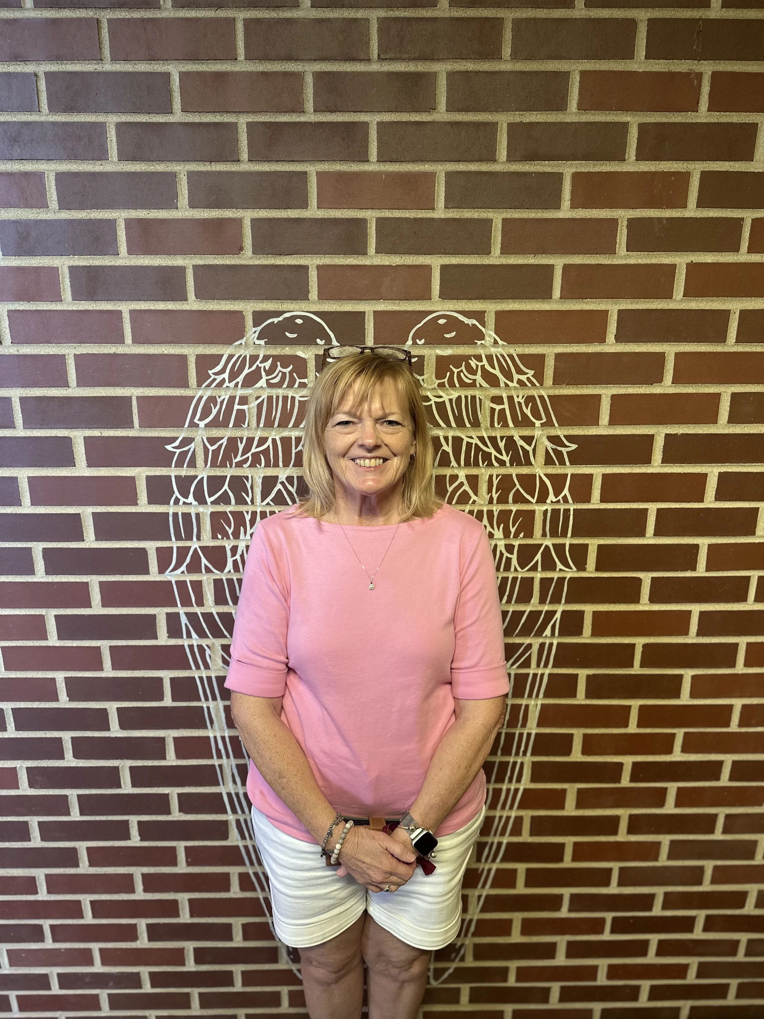 A middle-aged woman with blonde hair, wearing a pink long-sleeve shirt, white shorts, glasses on her head, and multiple bracelets, standing in front of a brick wall with a white outline of a dog on it, smiling at the camera.