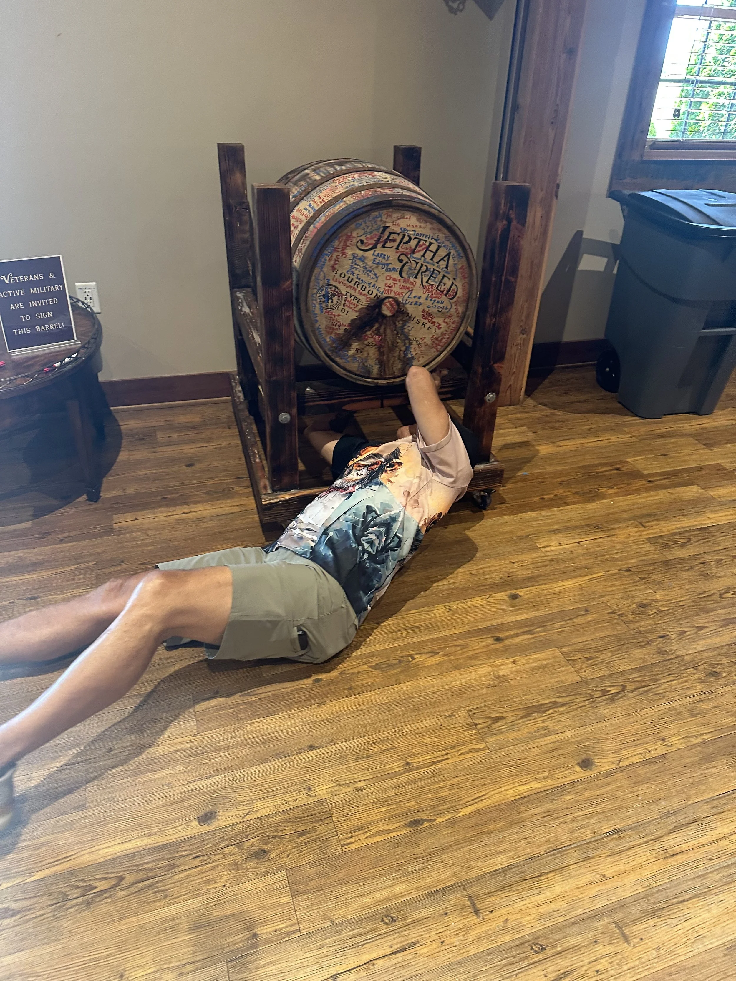 A person lying on the wooden floor underneath a large wooden barrel, which is held in a wooden frame. The barrel has writing and signatures on it. There is a sign on a table nearby that reads, 'Veterans & Active Military are invited to sign this barr