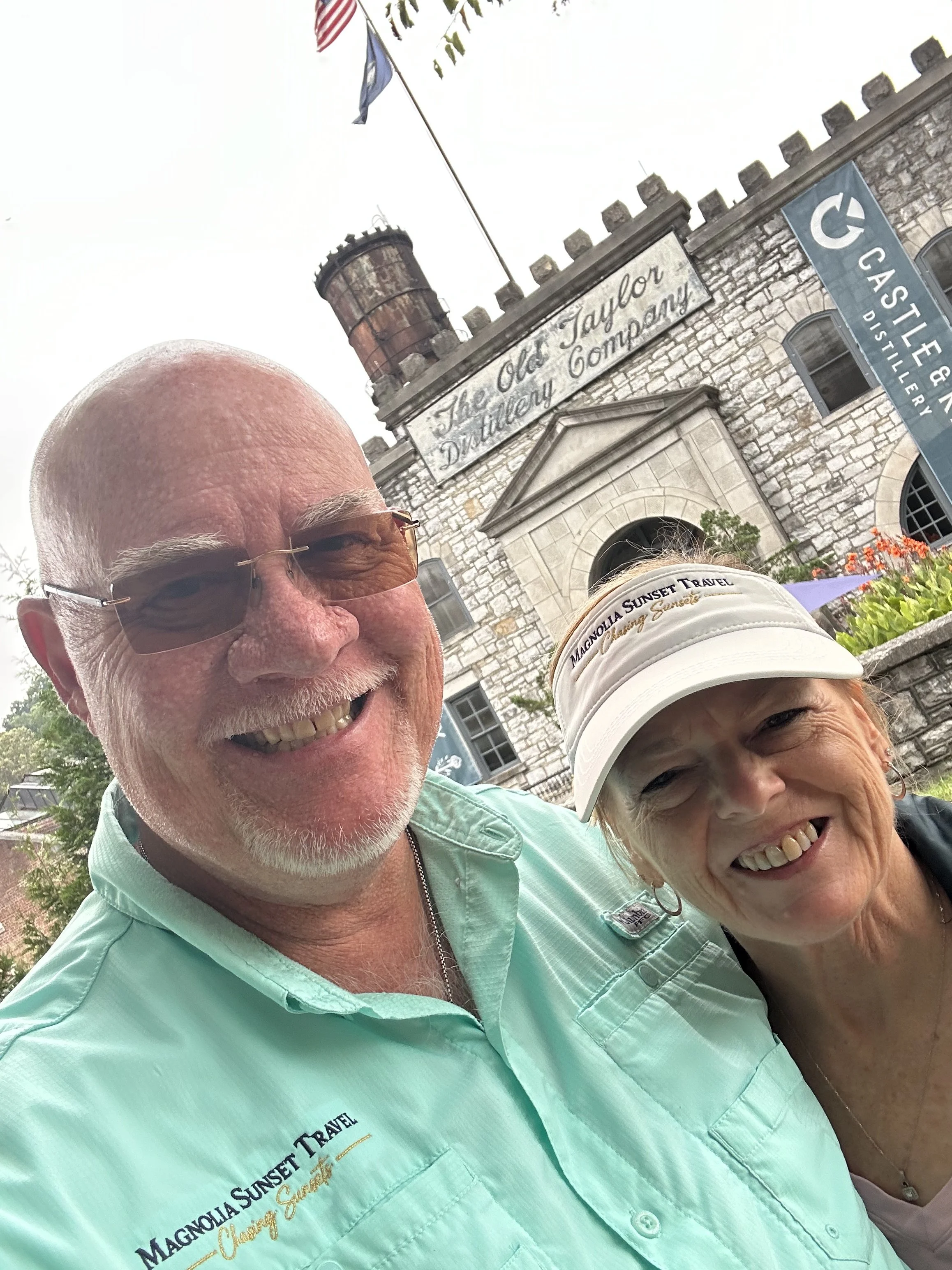 Close-up of a smiling older man wearing glasses and a light green shirt with logo and text, alongside a smiling older woman wearing a white cap with a logo, standing in front of a historic stone building with flags and a sign that reads "The Old Harb