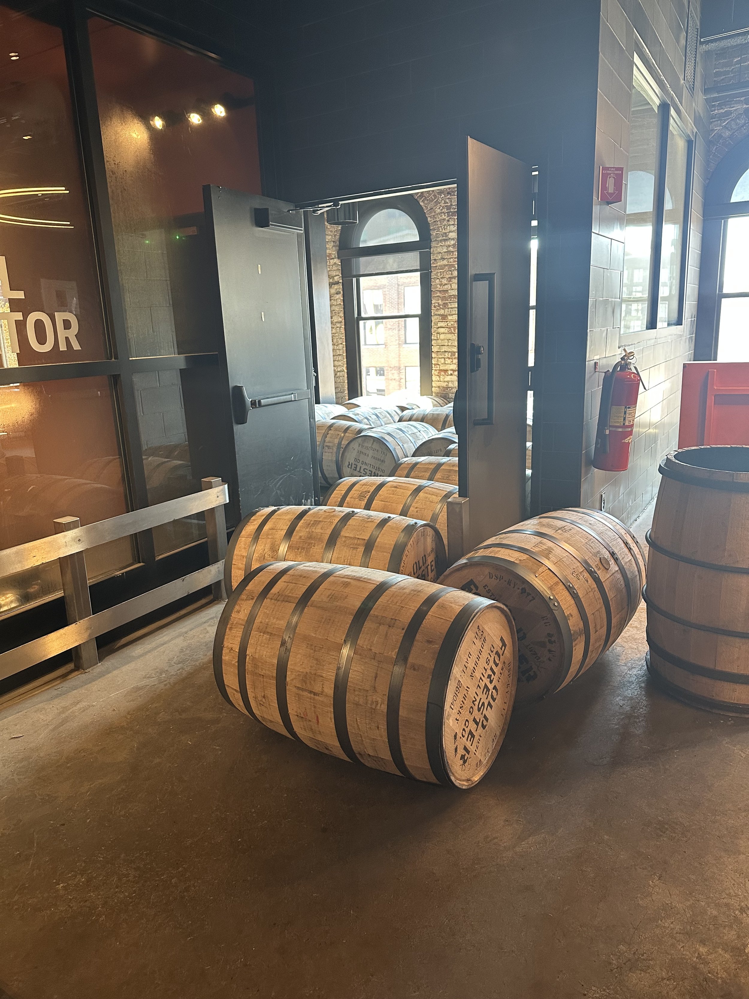 Wooden barrels stacked on the floor of a wine cellar or brewery, with a brick wall and large windows in the background.