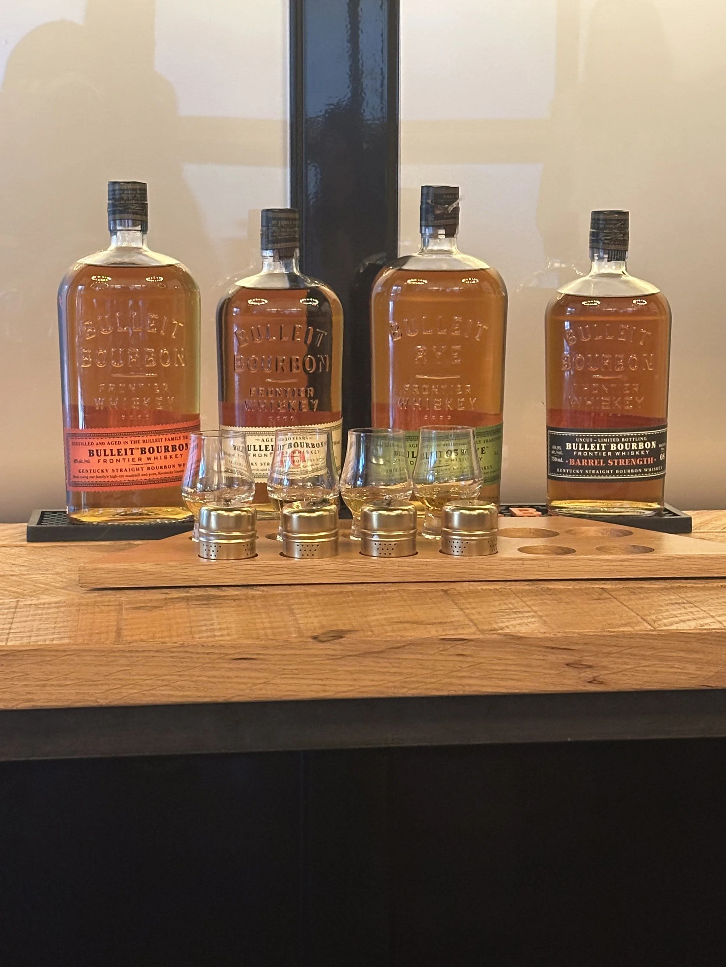 Four bottles of Bulleit Bourbon whiskey on a wooden surface, with four small glasses of whiskey in front of them, set against a wall with a black vertical stripe.