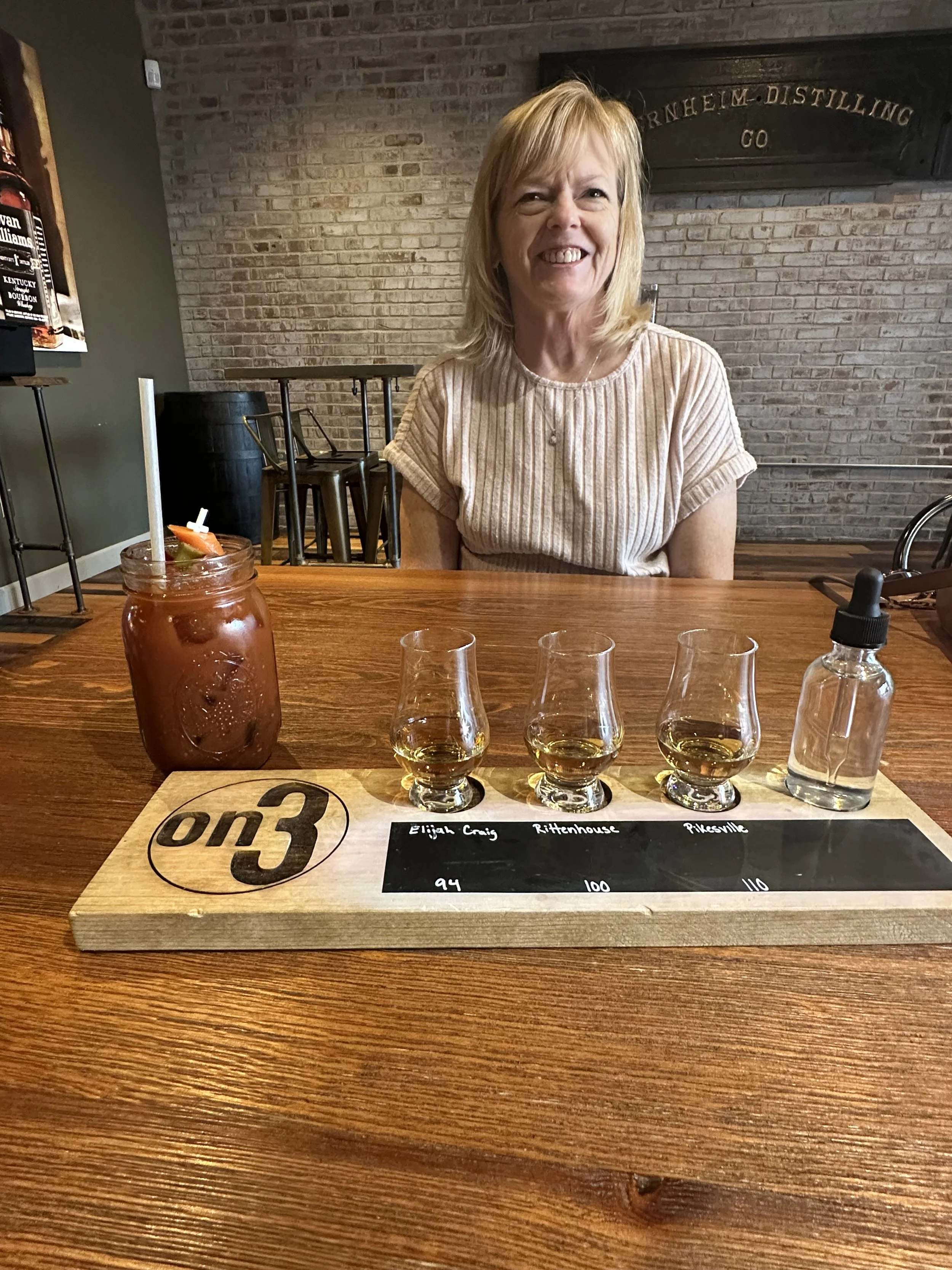 A woman is sitting at a wooden table with a flight of three whiskey glasses in front of her. She is smiling and wearing a beige striped shirt. Behind her is a brick wall with a sign that says "Furnheim Distilling Co." On the table is a wooden tasting