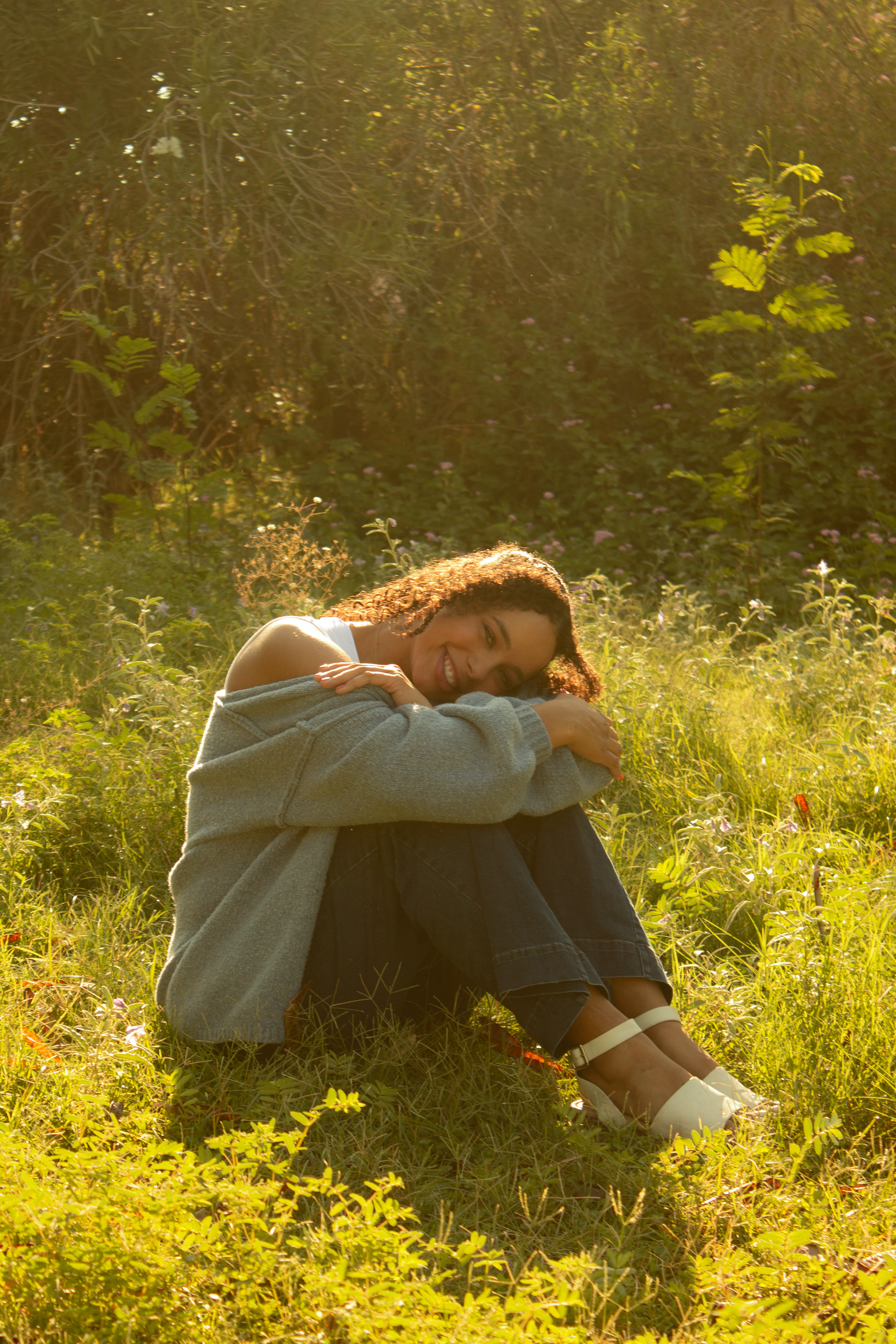 A trauma therapist with curly hair smiling as she sits on grass in a field, hugging her knees, with sunlight illuminating her face and a background of greenery and flowers.