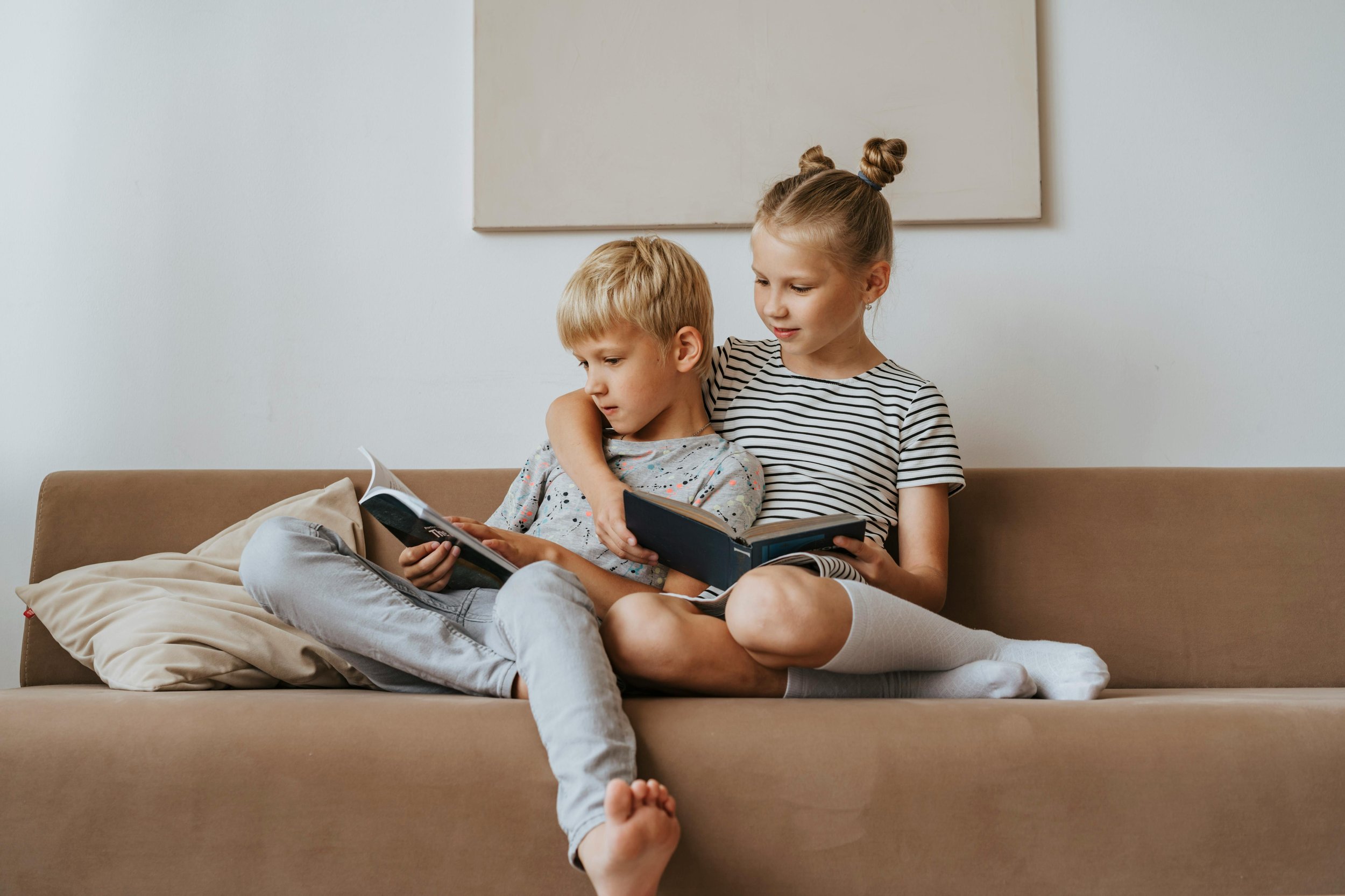 A young girl with her hair in buns and a boy with blonde hair sitting on a beige sofa, reading books together in a minimalistic room with white walls and a large canvas.