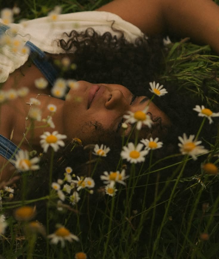 Person lying in a field of daisies with eyes closed, surrounded by green grass and flowers.