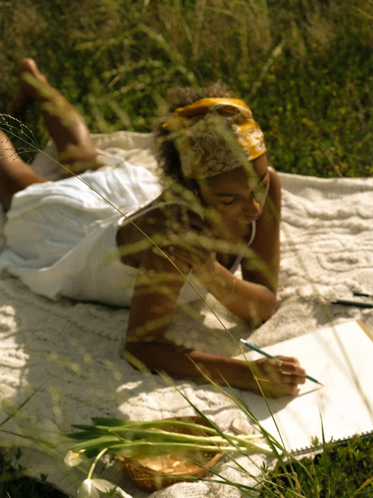 A woman lying on a blanket outdoors at night, writing or drawing on a paper with a pen, surrounded by grass and plants.