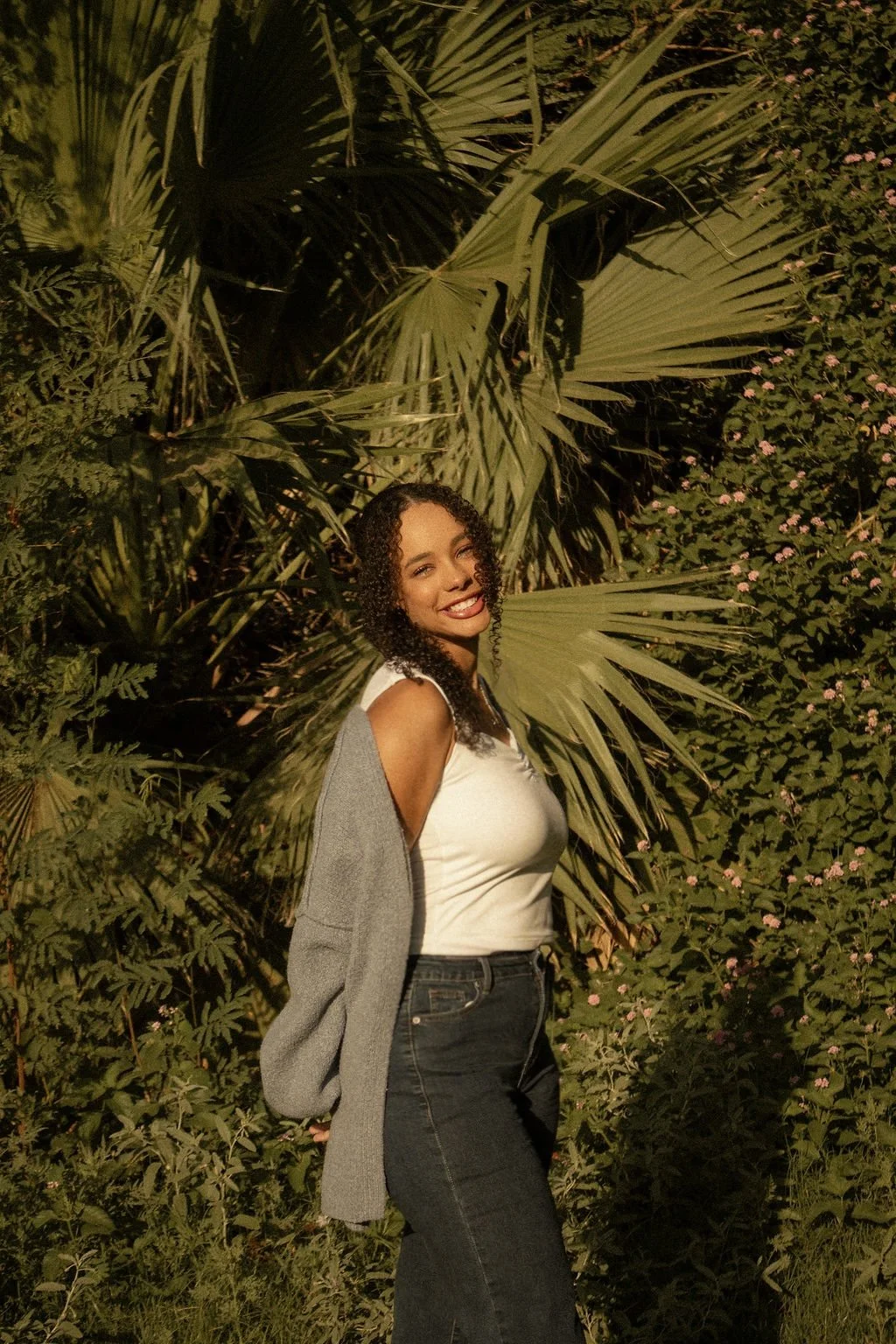 A young woman with curly dark hair smiling outdoors at night, standing in front of lush tropical plants and leaves with pink flowers.