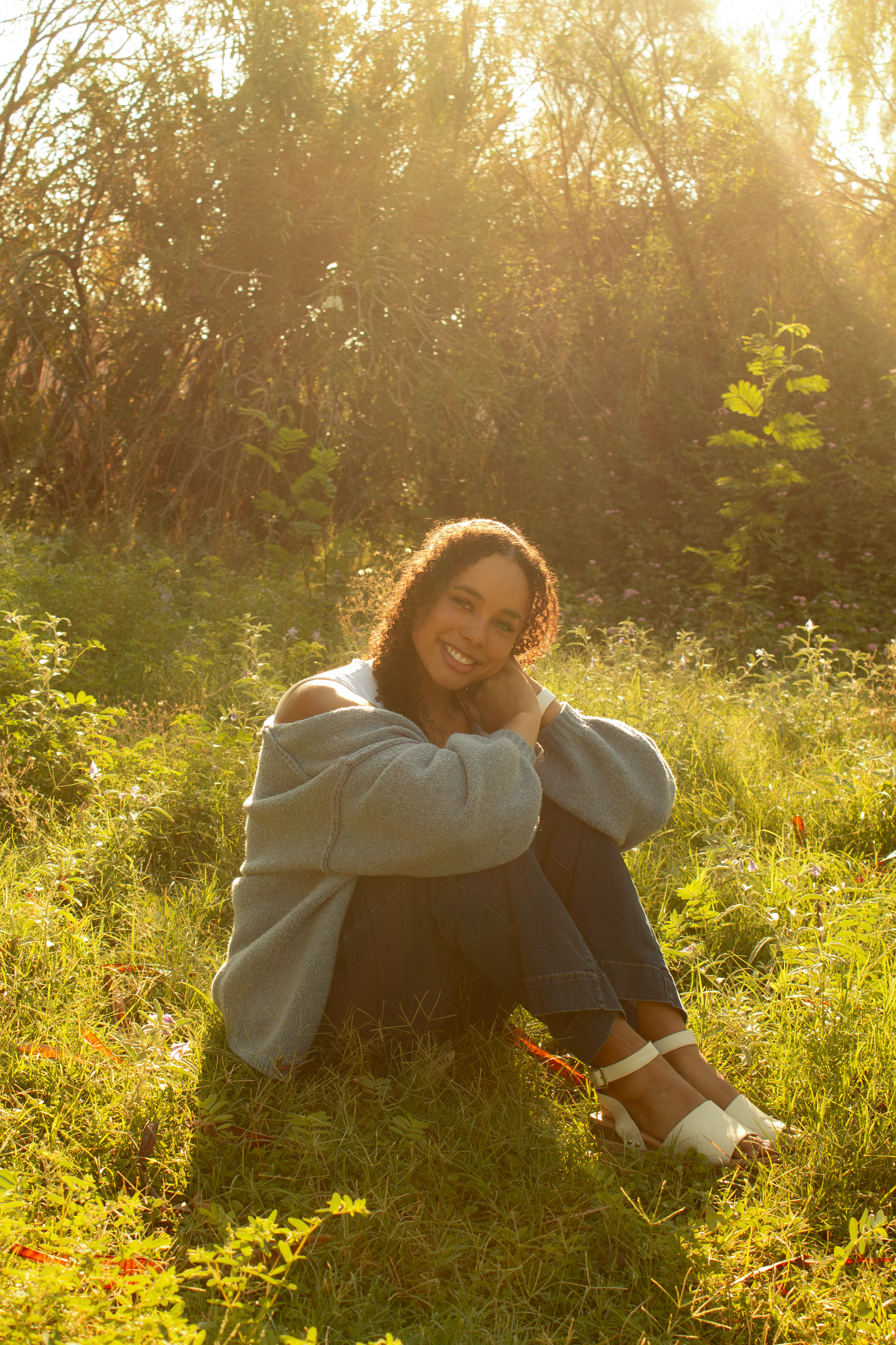 A woman with curly hair wearing a gray sweater and black pants sitting on grass in a field, smiling at the camera, with sunlight shining through trees in the background.